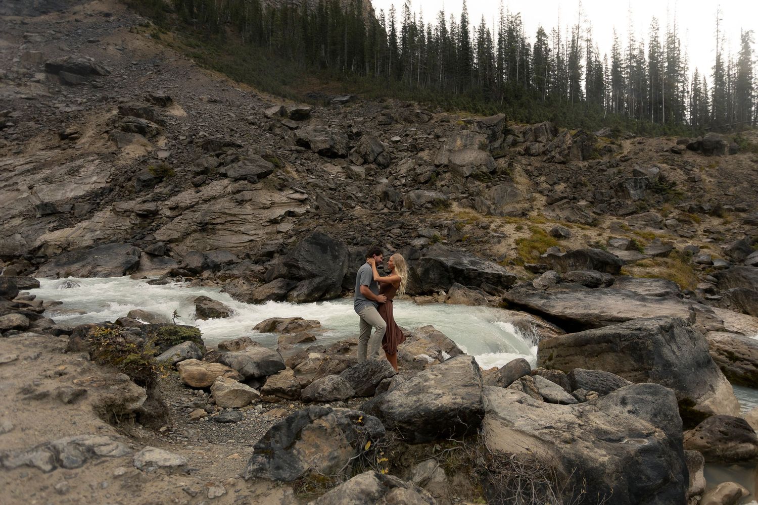 Two people embrace on rocky terrain with a mountain stream and evergreen forest in the background at Takakkaw Falls.
