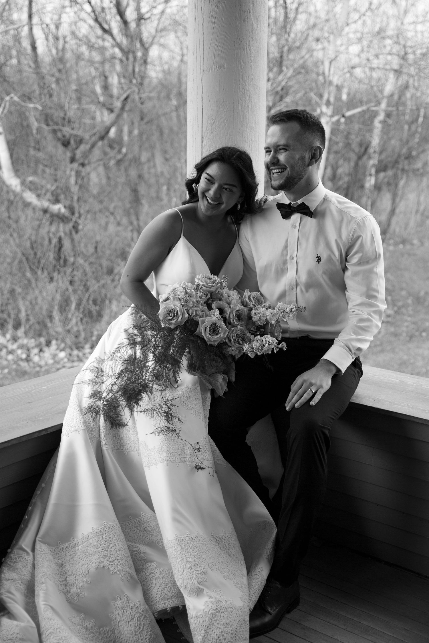 Black and white photo of a laughing bride holding a bouquet beside a smiling groom in a bow tie at their wedding.