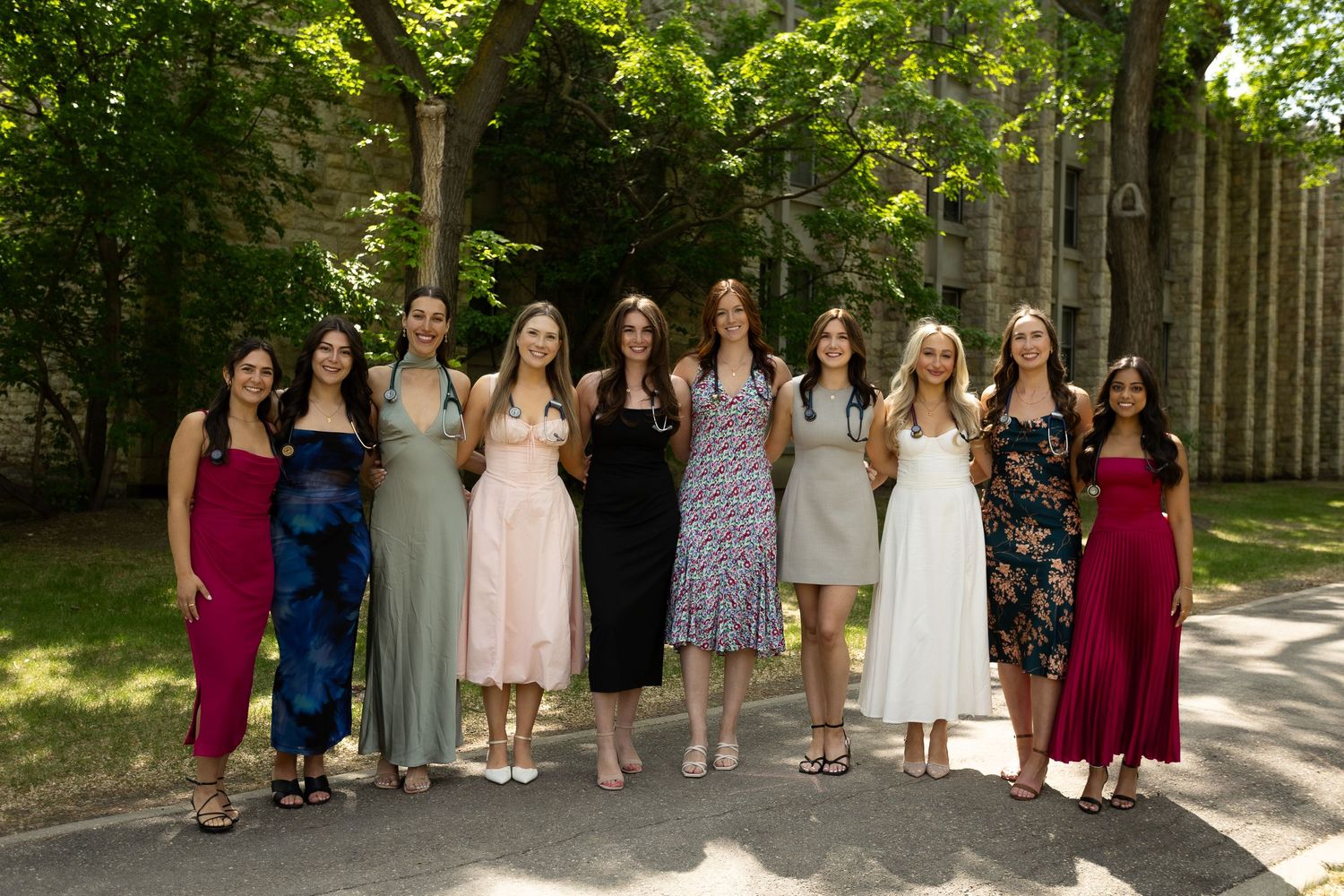 A group of ten women in elegant dresses posing together outdoors under lush green trees.