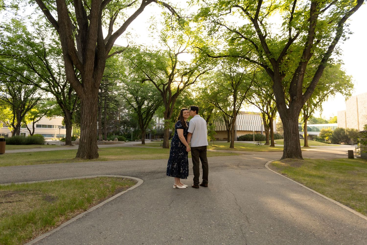 Couple holding hands on a curved park path surrounded by lush green trees during golden hour.