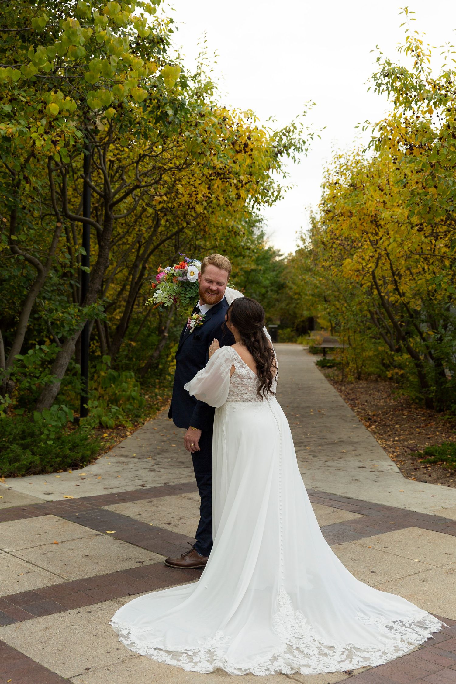 Bride and groom share a romantic moment on a brick path surrounded by golden autumn trees, bride in lace gown with long train.