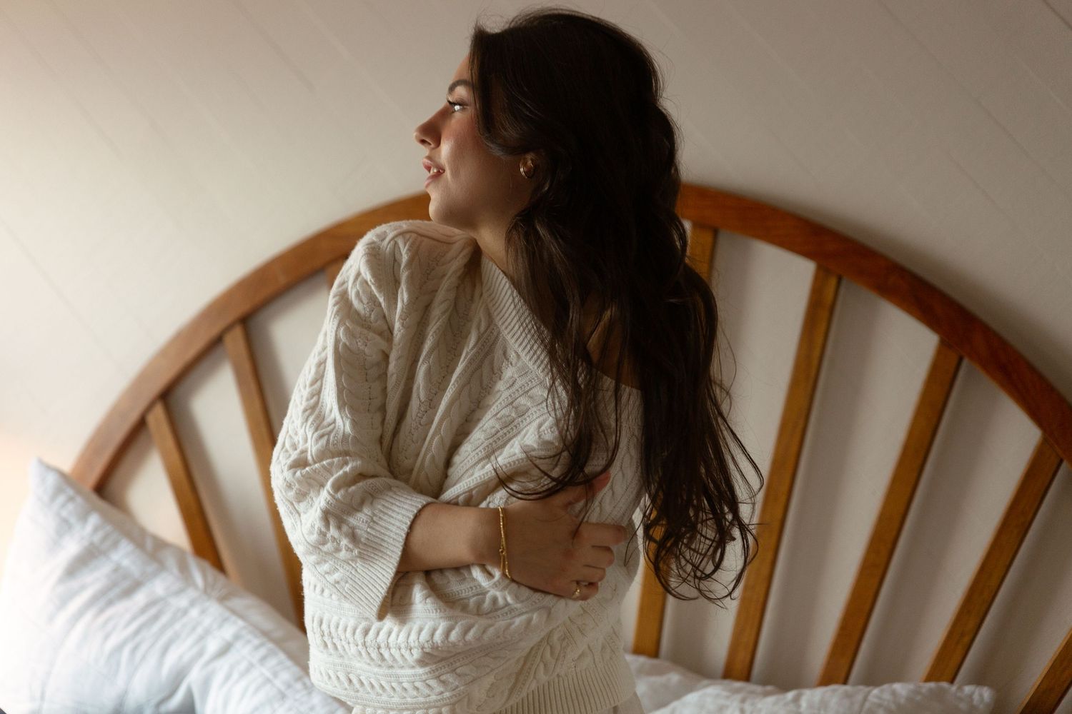 A young woman with long dark hair sits on a bed in a cozy cream knit sweater, gazing thoughtfully to the side.