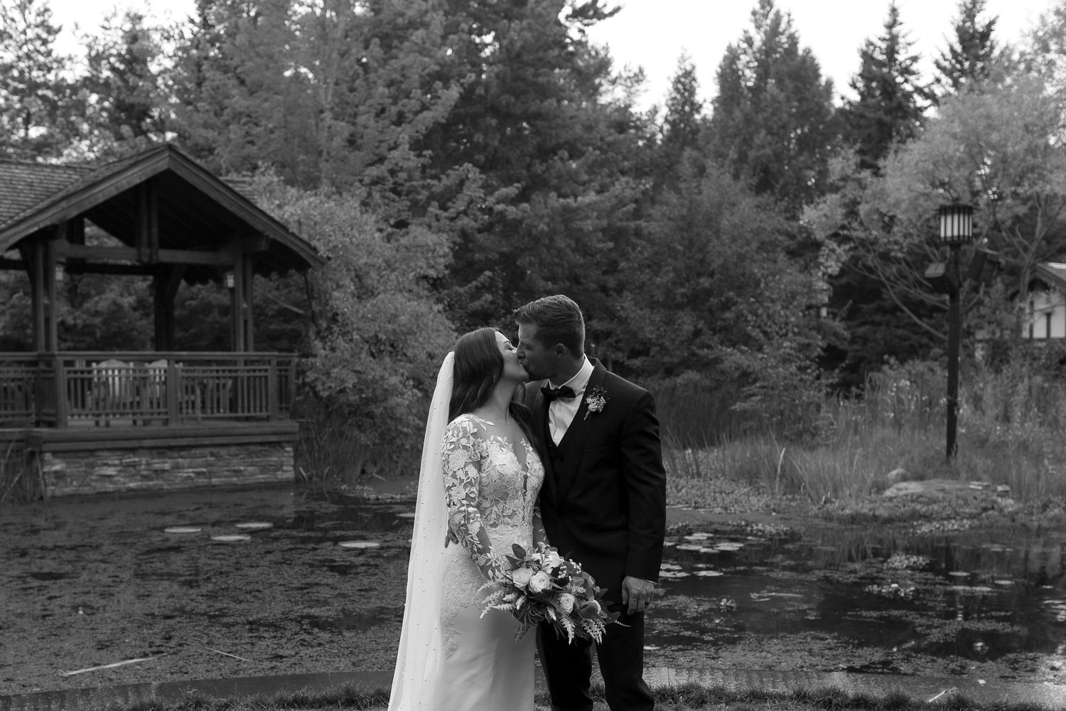 Black and white photo of a bride and groom sharing a kiss outdoors near a rustic log cabin surrounded by tall trees.