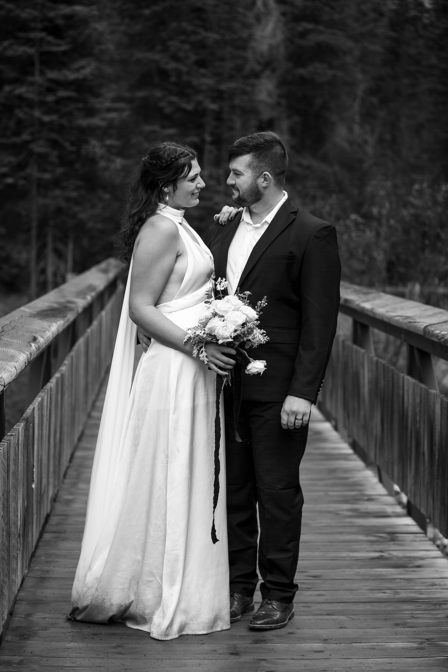 A black and white wedding photo of a couple standing together on a wooden bridge surrounded by evergreen trees.