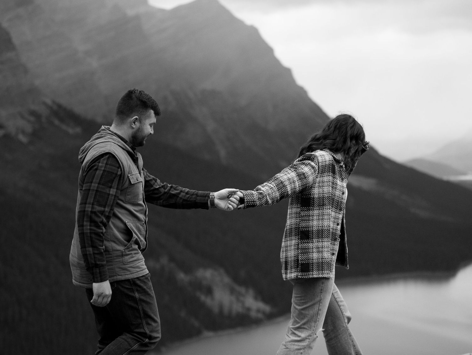 Black and white photo of a couple holding hands on a mountain overlook with a lake and peaks in the background.
