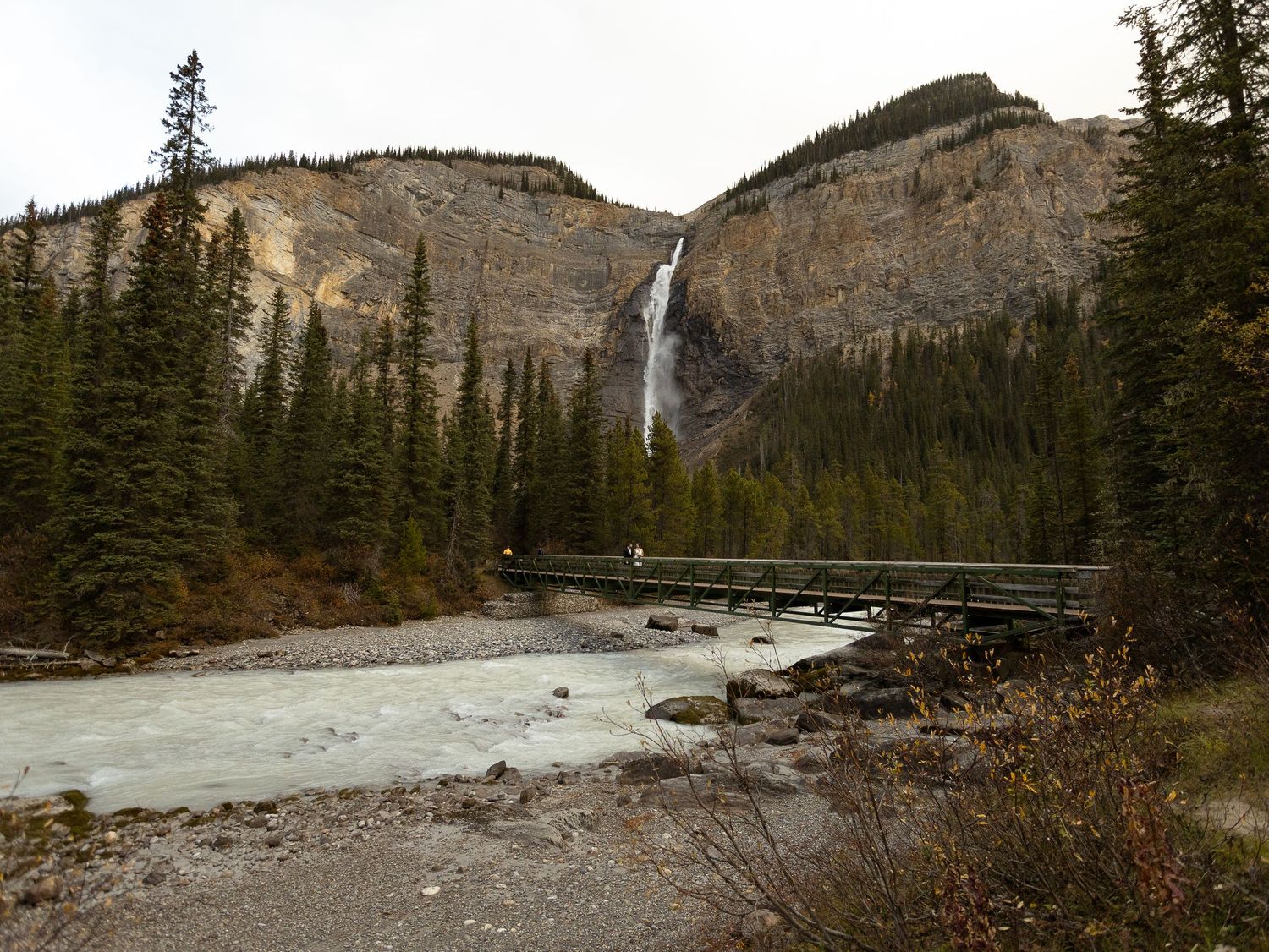 Takakkaw Falls cascading down rocky cliffs with a wooden bridge over a turquoise river surrounded by evergreen trees.