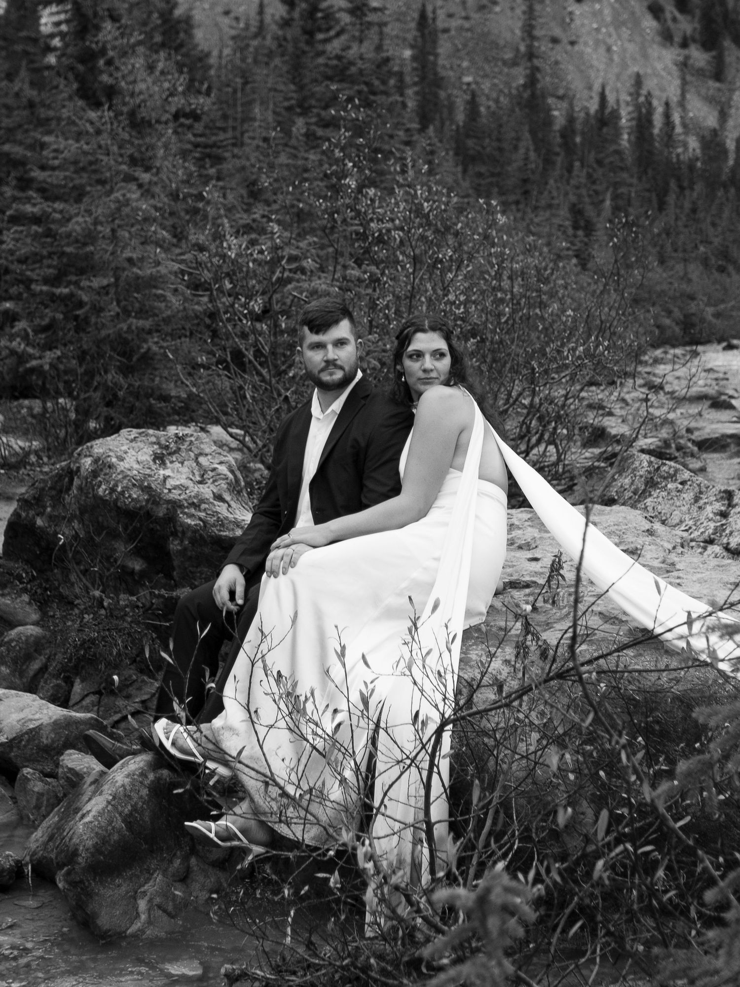 Black and white photo of a bride and groom sitting on rocks in a mountain forest setting.