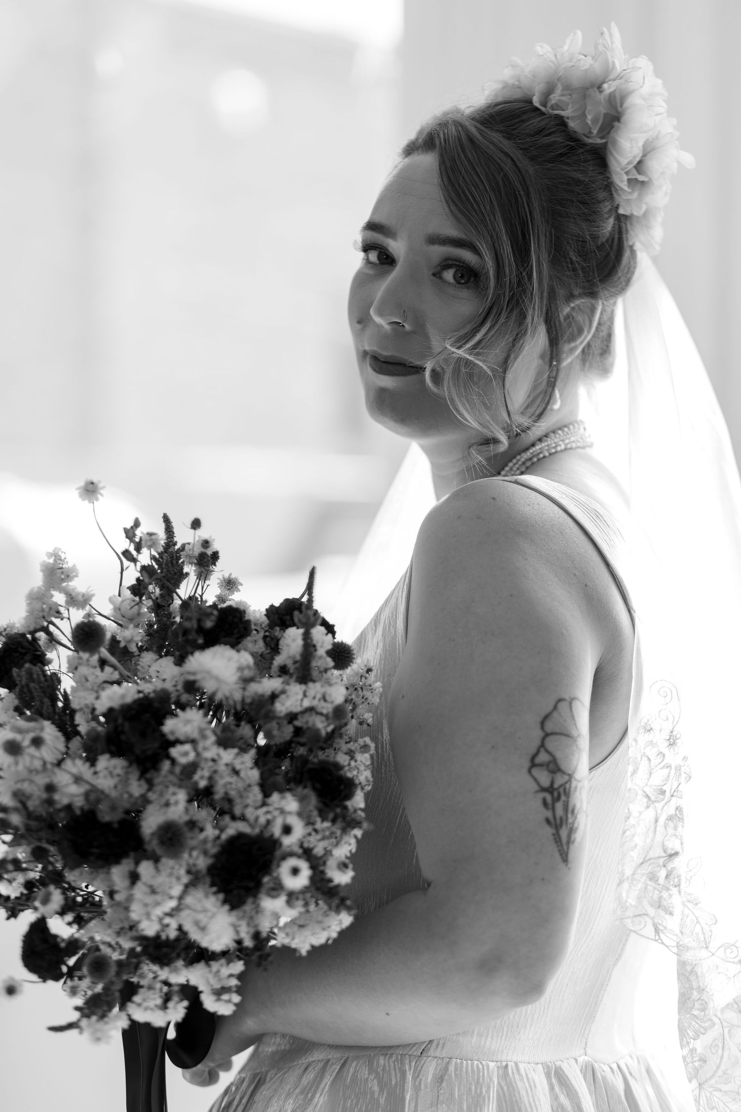 Black and white portrait of a bride holding a bouquet, wearing a veil and floral hair accessory, with a tattoo on her arm.