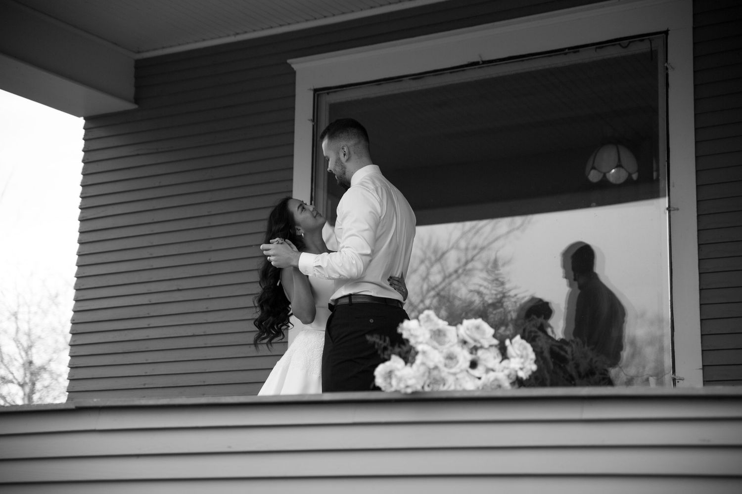 Black and white photo of a bride and groom sharing a romantic embrace on a porch with white flowers nearby.