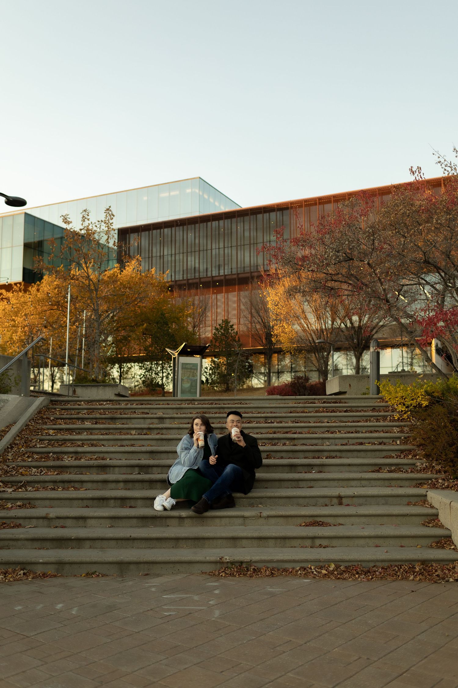 Couple sitting on outdoor steps of a modern building surrounded by autumn foliage on a clear day.