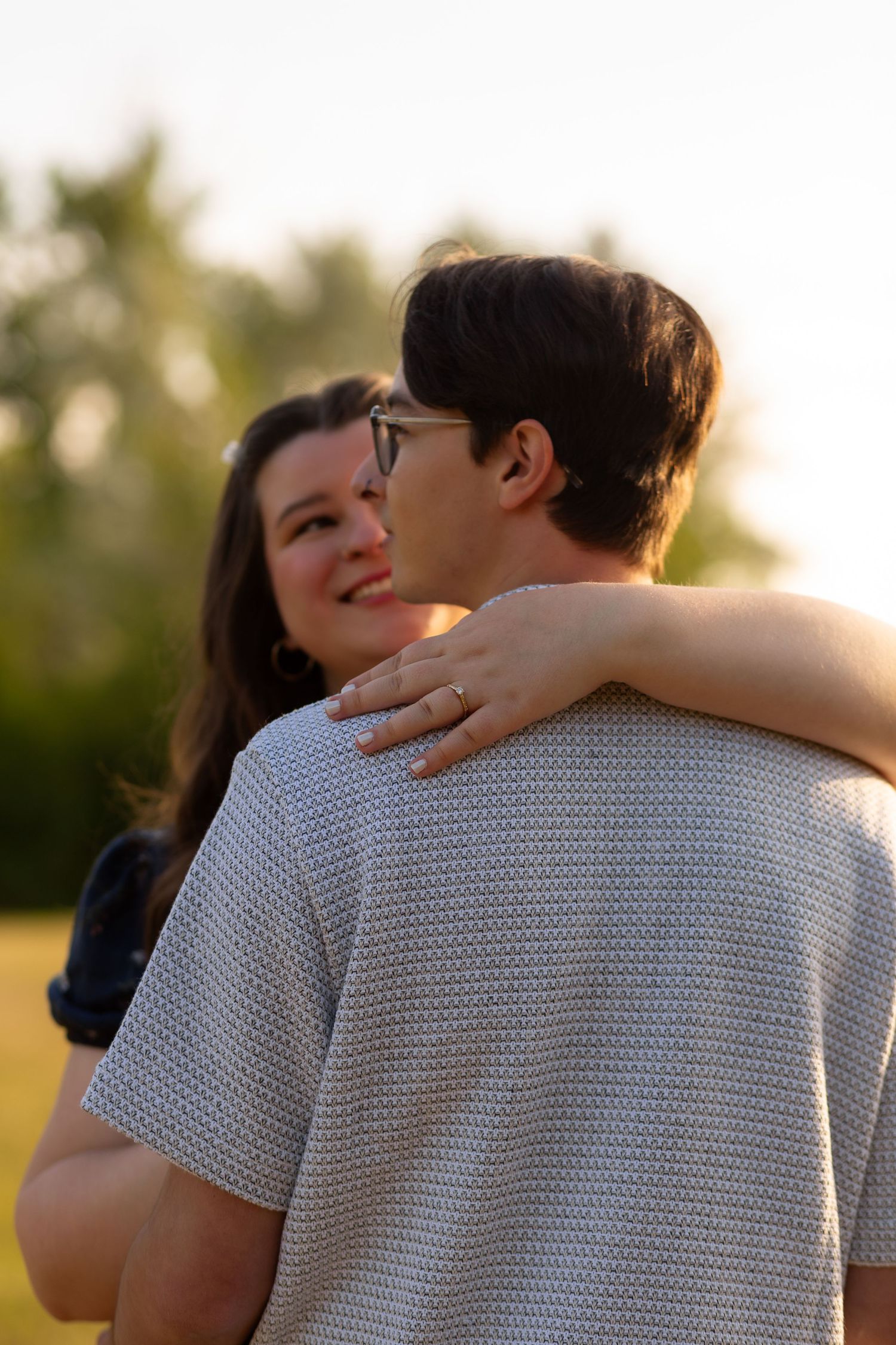 A couple embraces outdoors at golden hour, woman smiling and showing engagement ring on man's back.