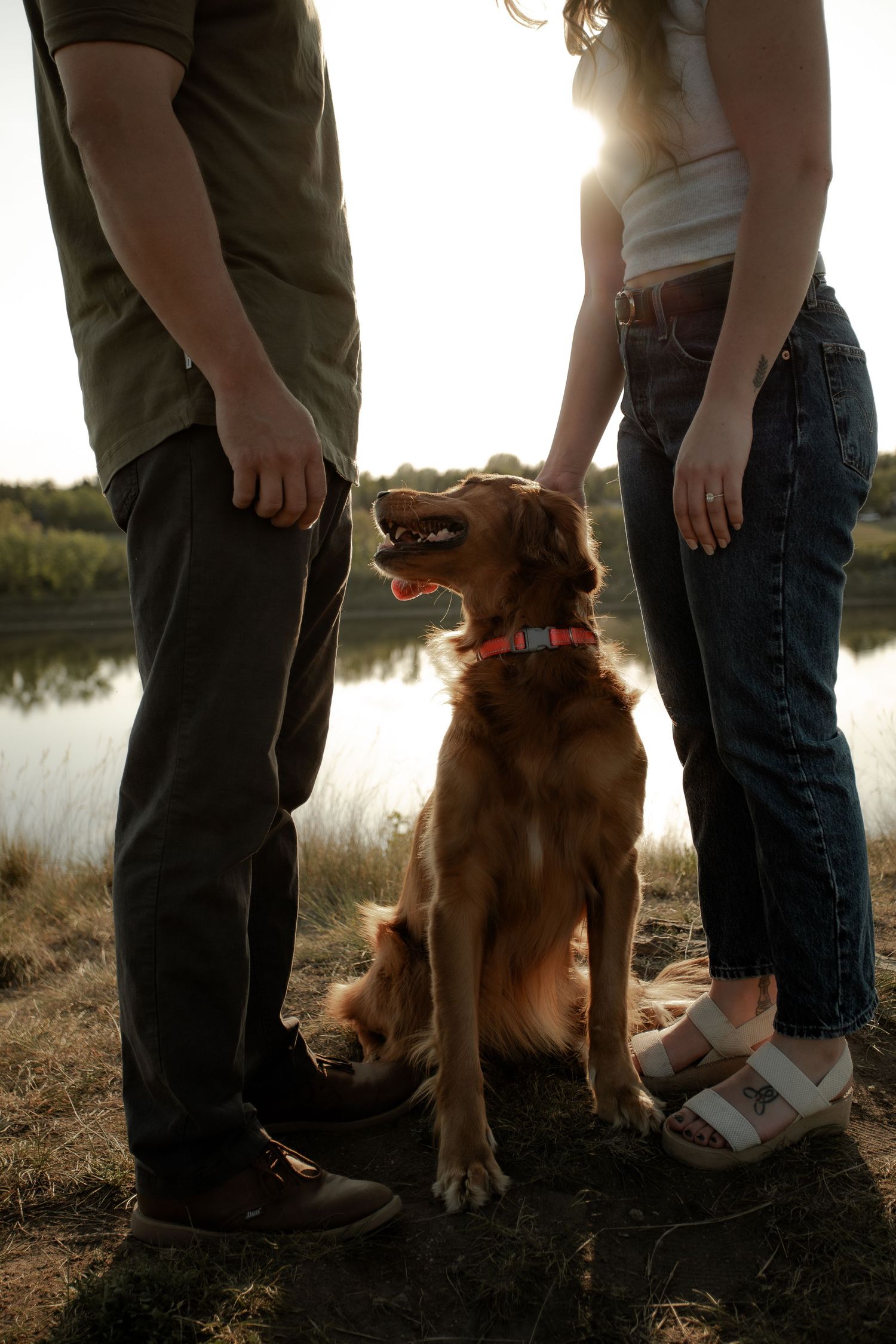 A golden retriever sits between two people's legs during sunset by a lake.