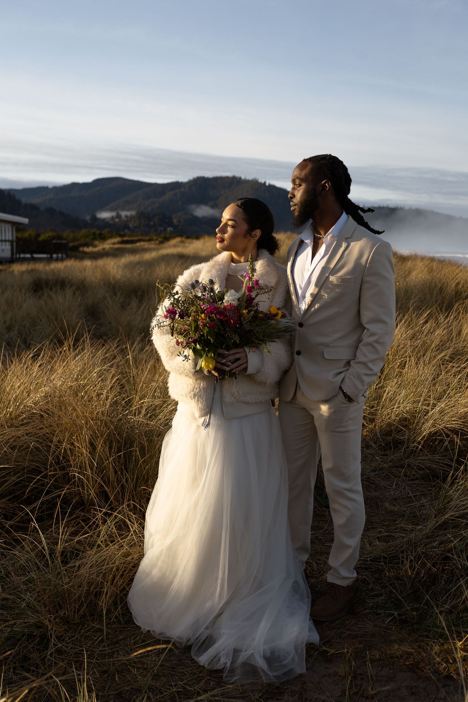 Bride and groom in wedding attire stand in golden grass field with mountain backdrop at sunset.