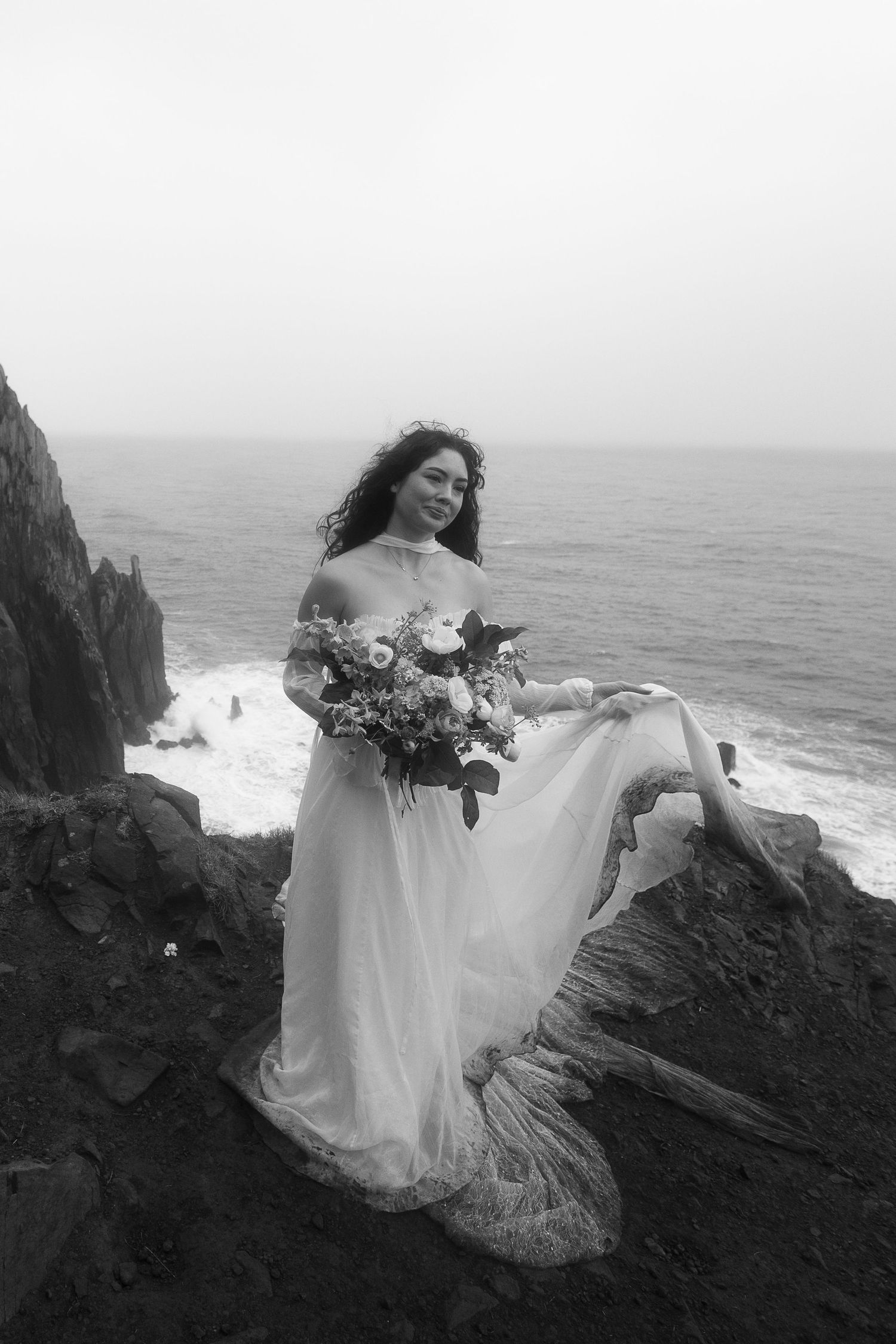 Black and white photo of a bride in flowing gown holding bouquet on rocky ocean cliffside.