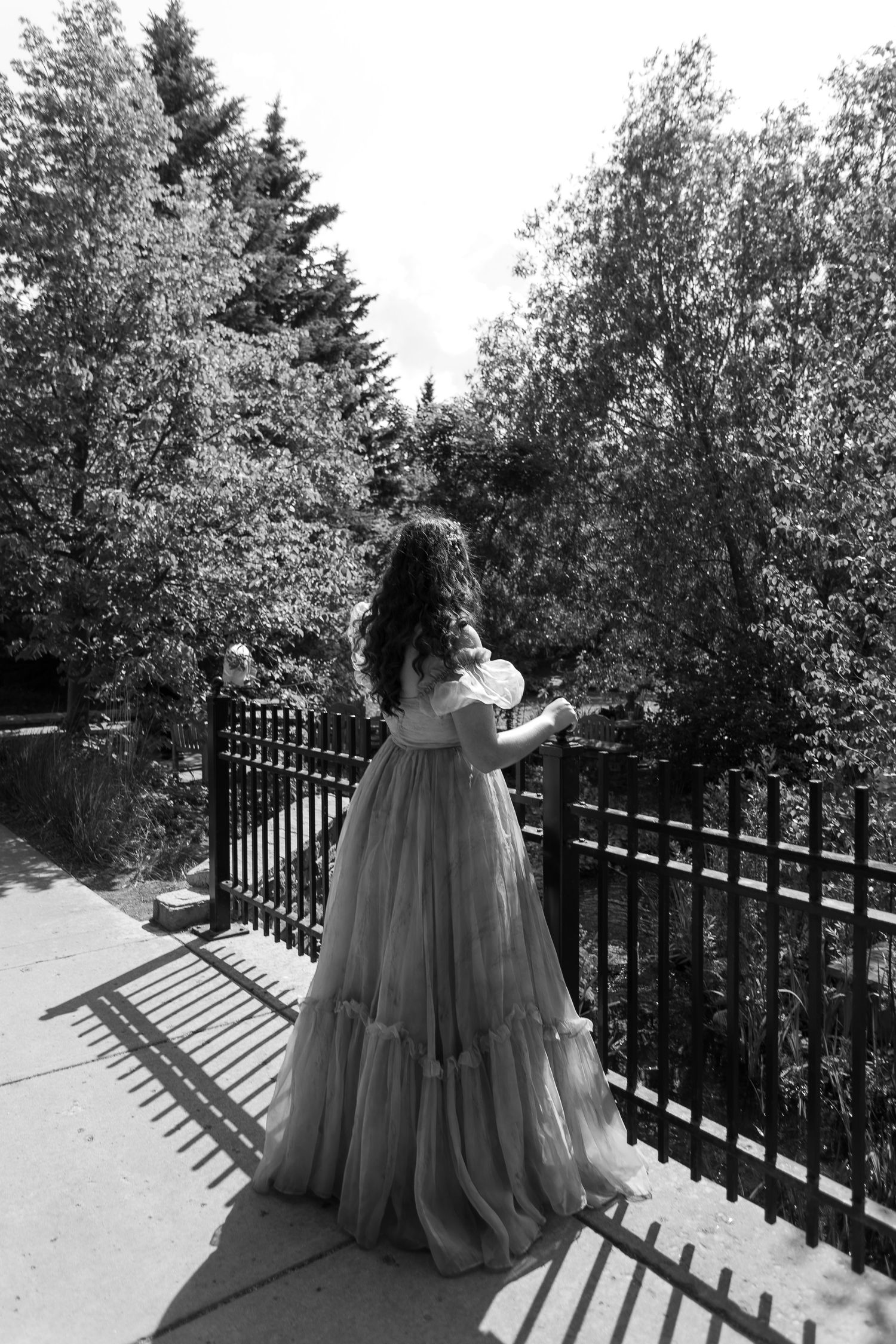 Black and white photo of a woman in a flowing ball gown standing on a sunlit path by iron railings surrounded by trees.