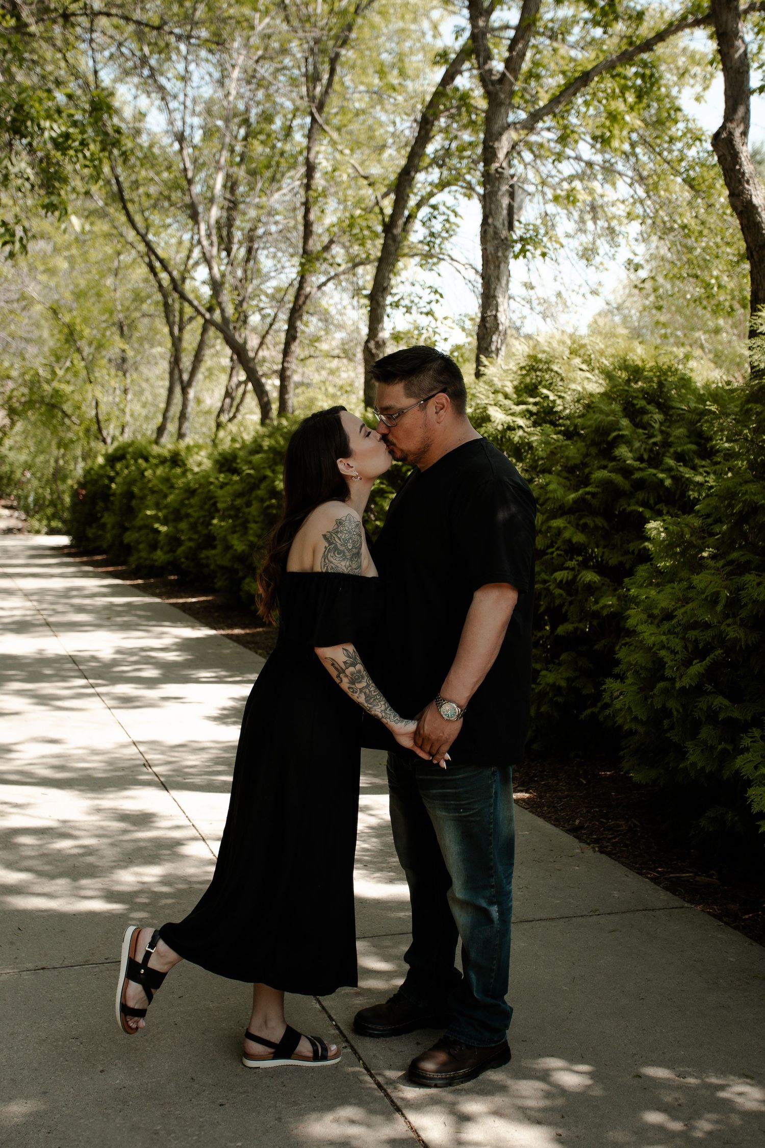 Couple kissing on a sunny garden path, woman in black dress with tattoos, man in black shirt and jeans.