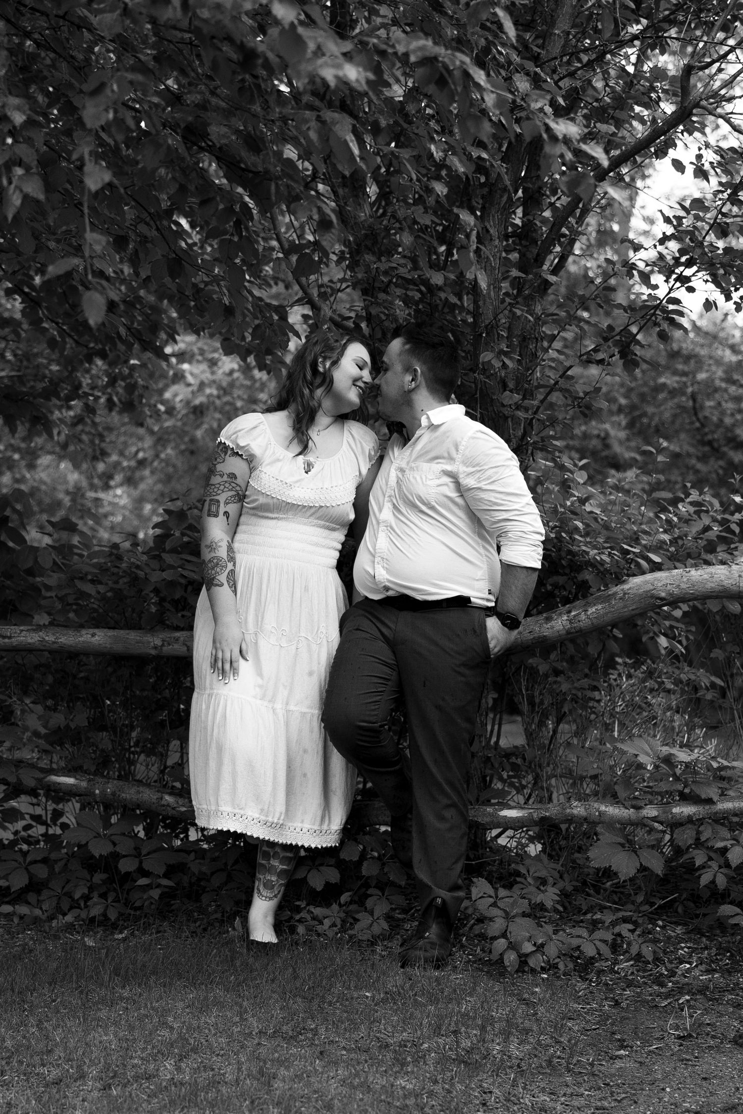 Black and white photo of a couple sharing a romantic moment by a rustic fence surrounded by lush greenery.
