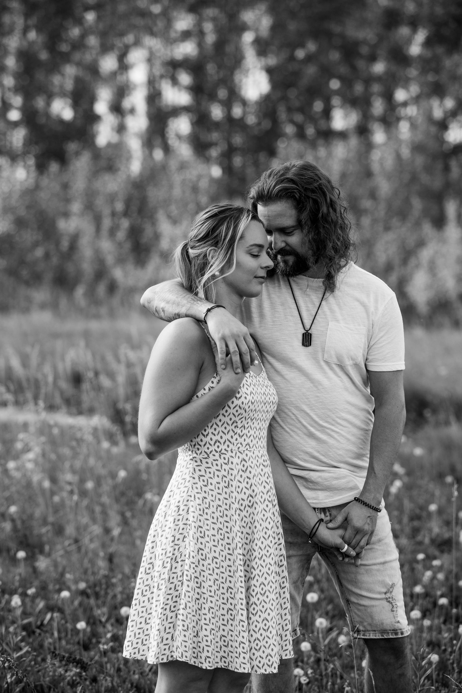 Black and white photo of a romantic couple embracing in a field, man with curly hair and woman in summer dress.