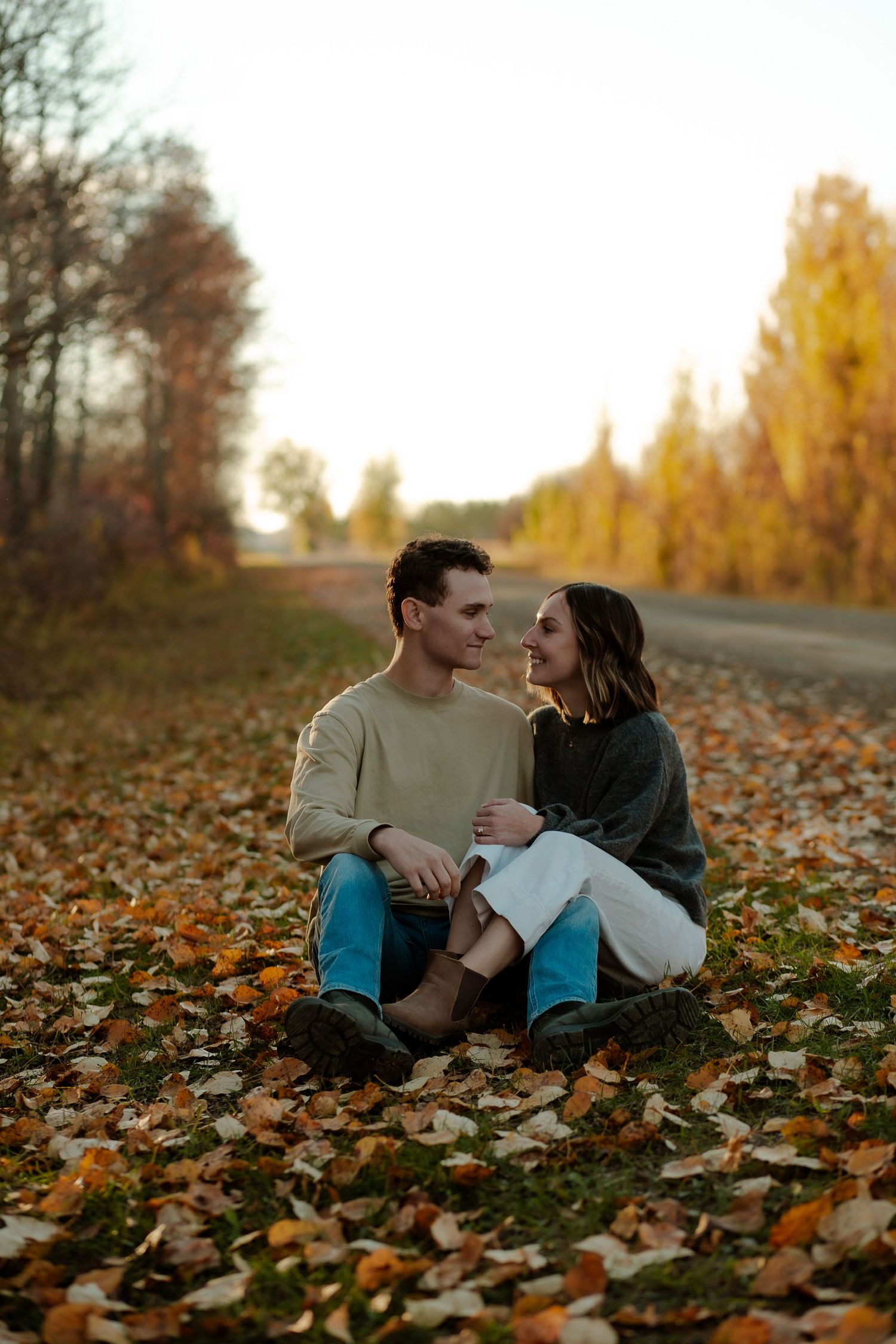 Couple sitting together on fallen autumn leaves beside a scenic road surrounded by colorful fall foliage at sunset.