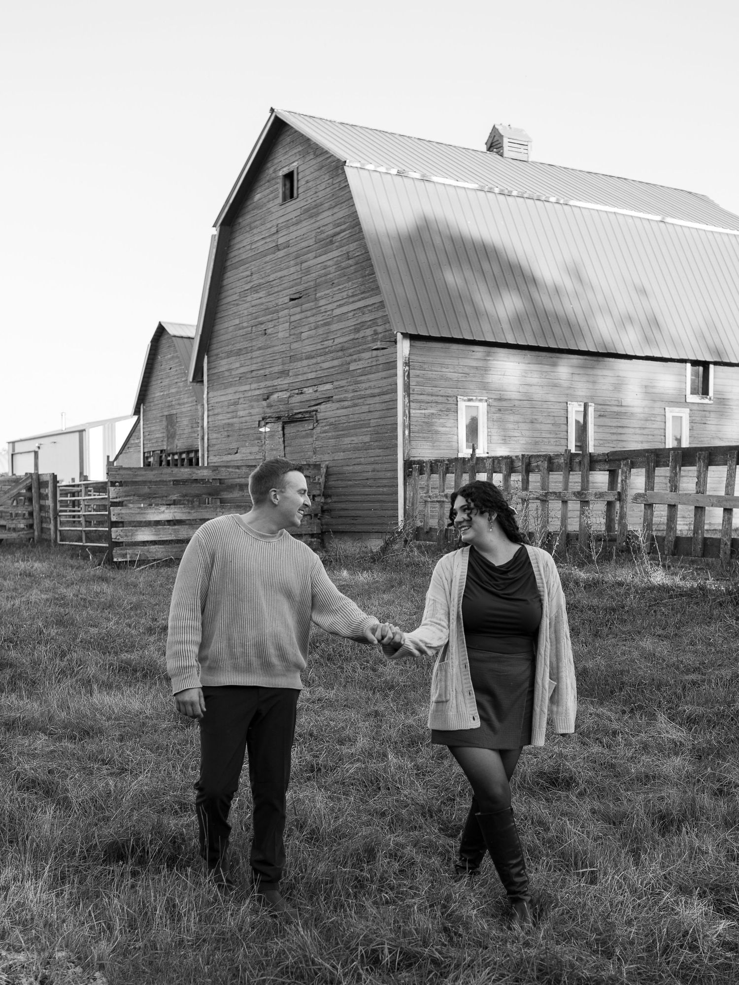 Two people walk hand in hand across a field with rustic wooden barns in the background on a prairie farm.