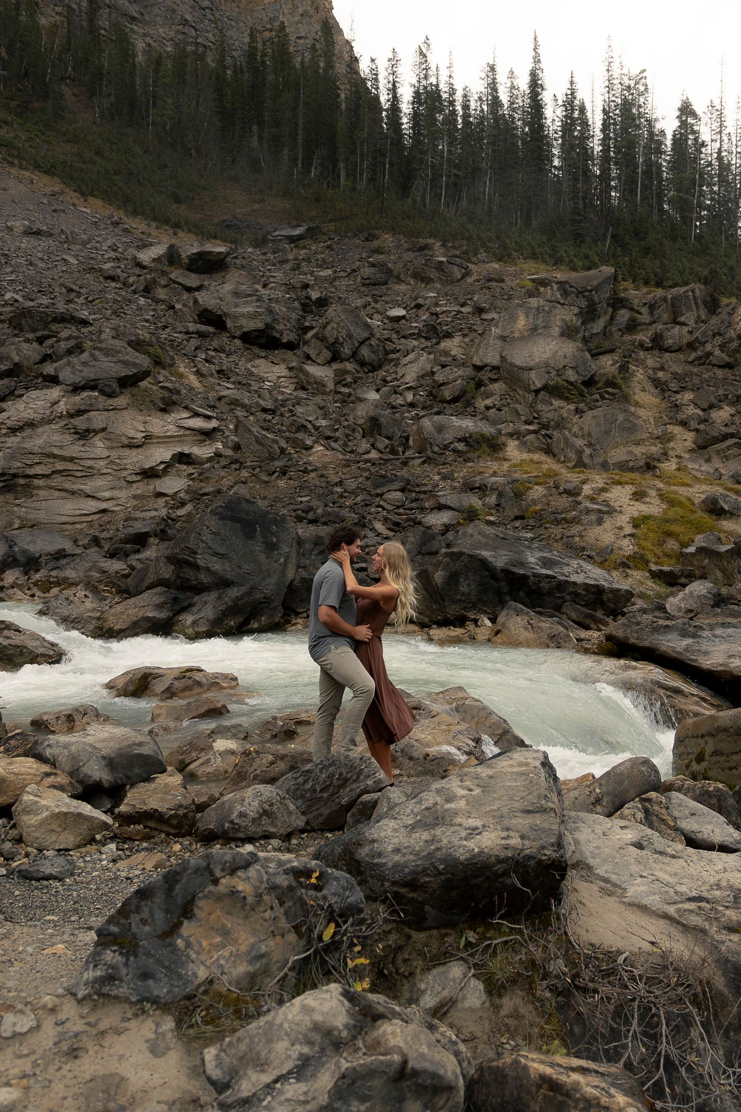 A couple shares an intimate moment while standing on rocky terrain near rushing water and pine trees at Takakkaw Falls in British Colombia.