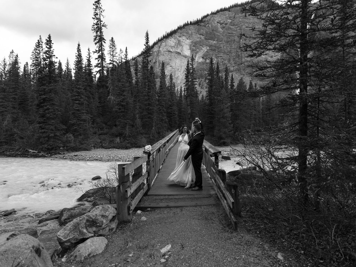 Black and white photo of a couple in wedding attire kissing on a wooden bridge surrounded by pine trees and a mountain river.