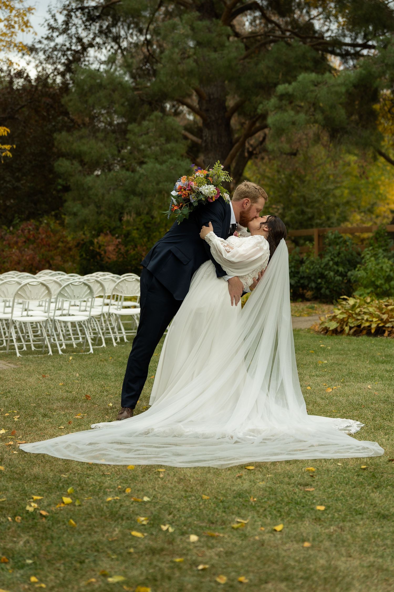 A wedding couple poses dramatically on grass with white chairs in background at the Forestry Farm in Saskatoon.
