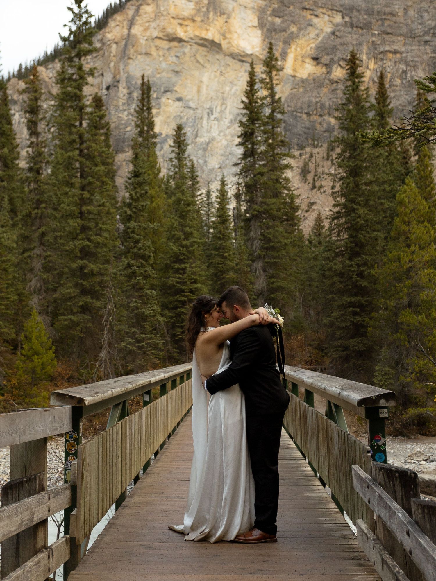 Bride and groom share a romantic kiss on a wooden bridge surrounded by evergreen trees and rocky mountains.