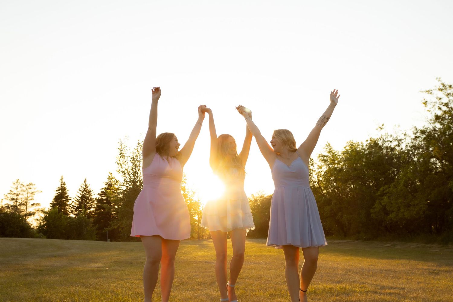Three women in dresses celebrate at sunset, raising their arms joyfully on a green lawn with trees in the background.