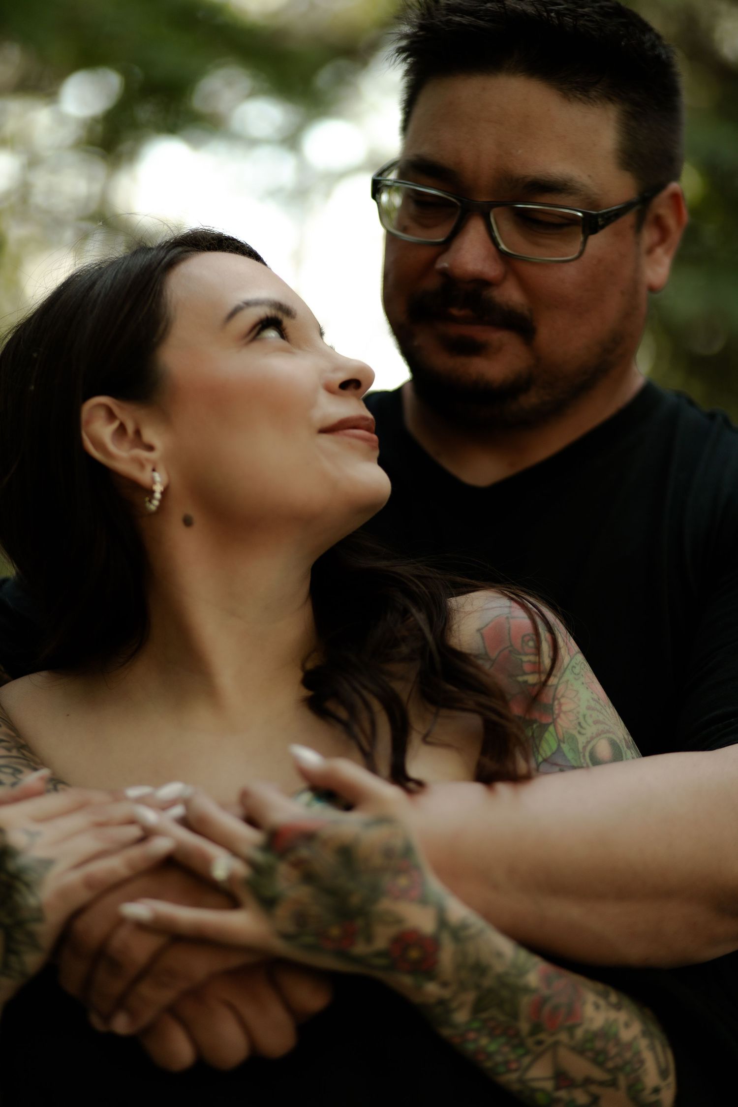 A tattooed couple embraces outdoors, the woman gazing up lovingly while the man holds her hands around her waist.