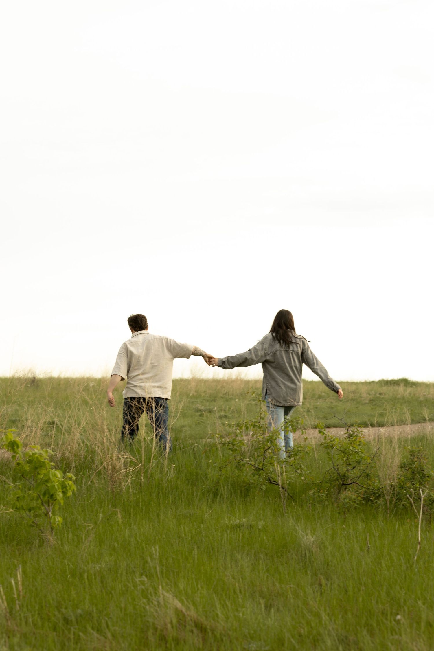 A couple holds hands while walking through a lush green meadow, viewed from behind on a cloudy day.