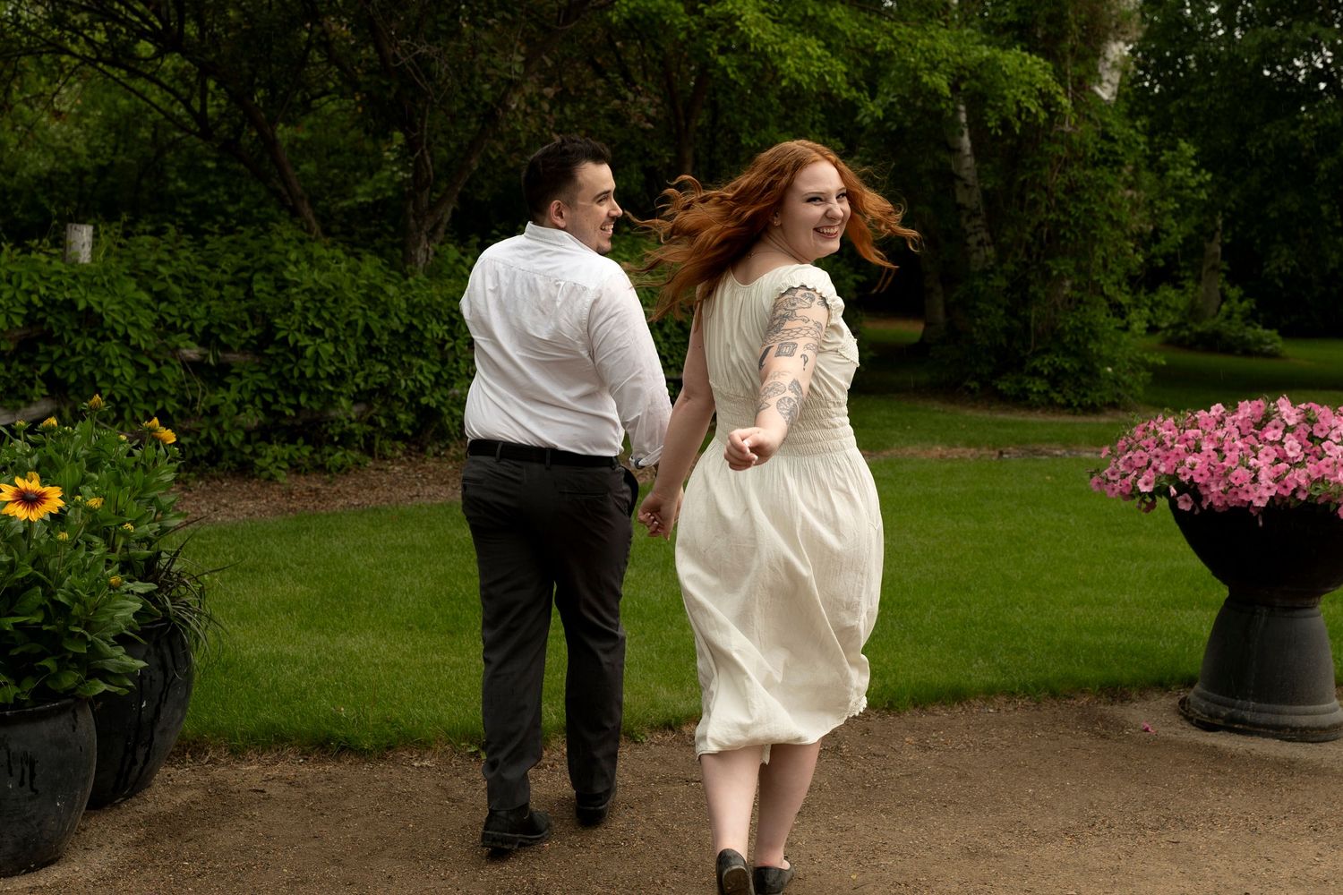 Couple in wedding attire walking hand-in-hand through a lush garden, woman in white dress with flowing red hair.