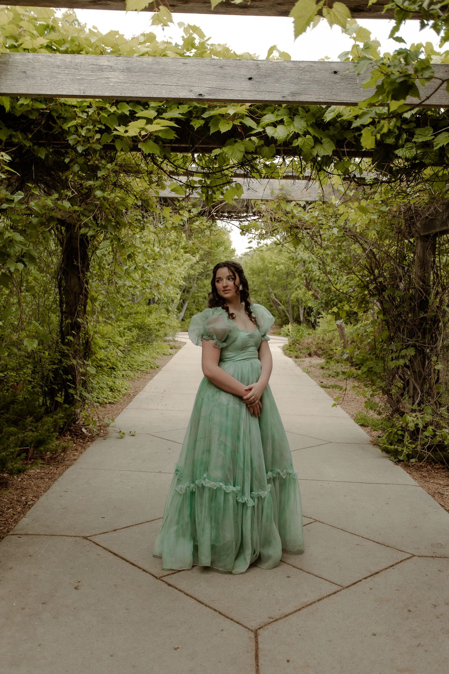 Woman in mint green tulle ball gown standing under vine-covered pergola in lush garden pathway.