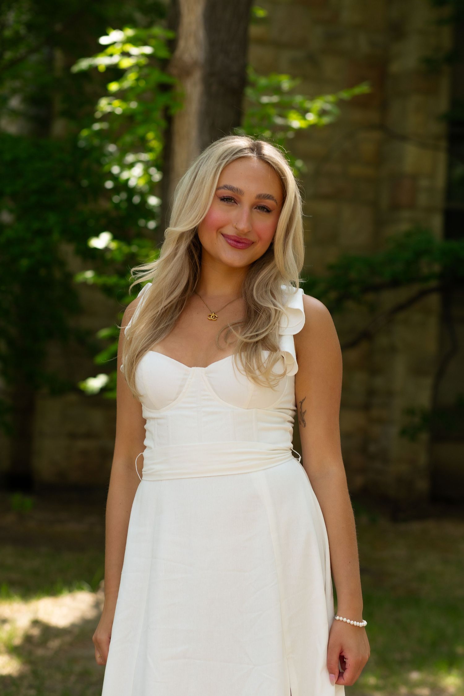 Young blonde woman smiling in white one-shoulder dress, posing outdoors among green trees and stone wall.