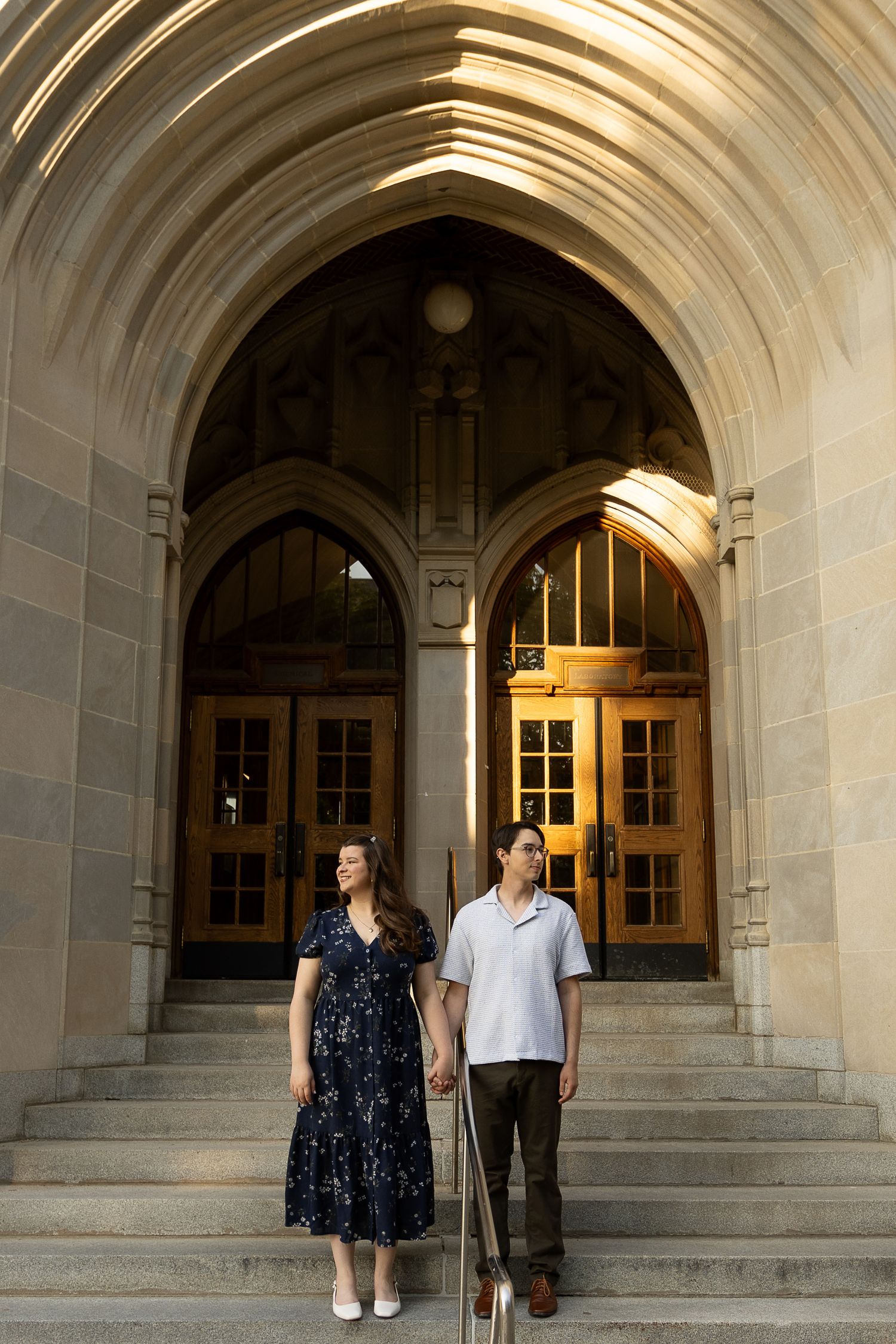 Couple holding hands on stone steps beneath ornate Gothic arch entrance of historic building.
