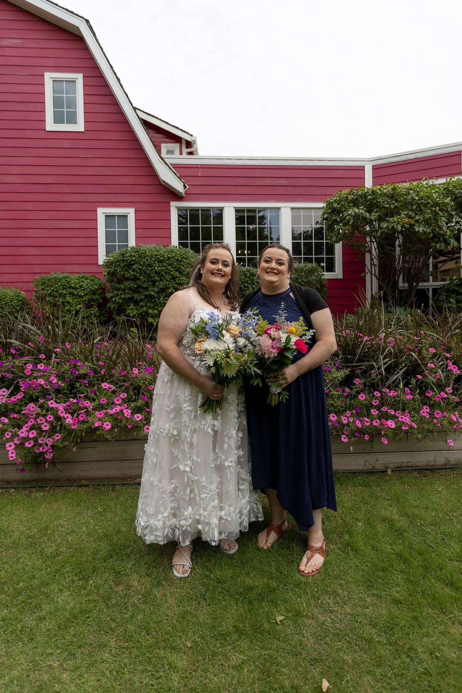 Two women in formal dresses holding bouquets stand in front of a red barn with colorful flowers.