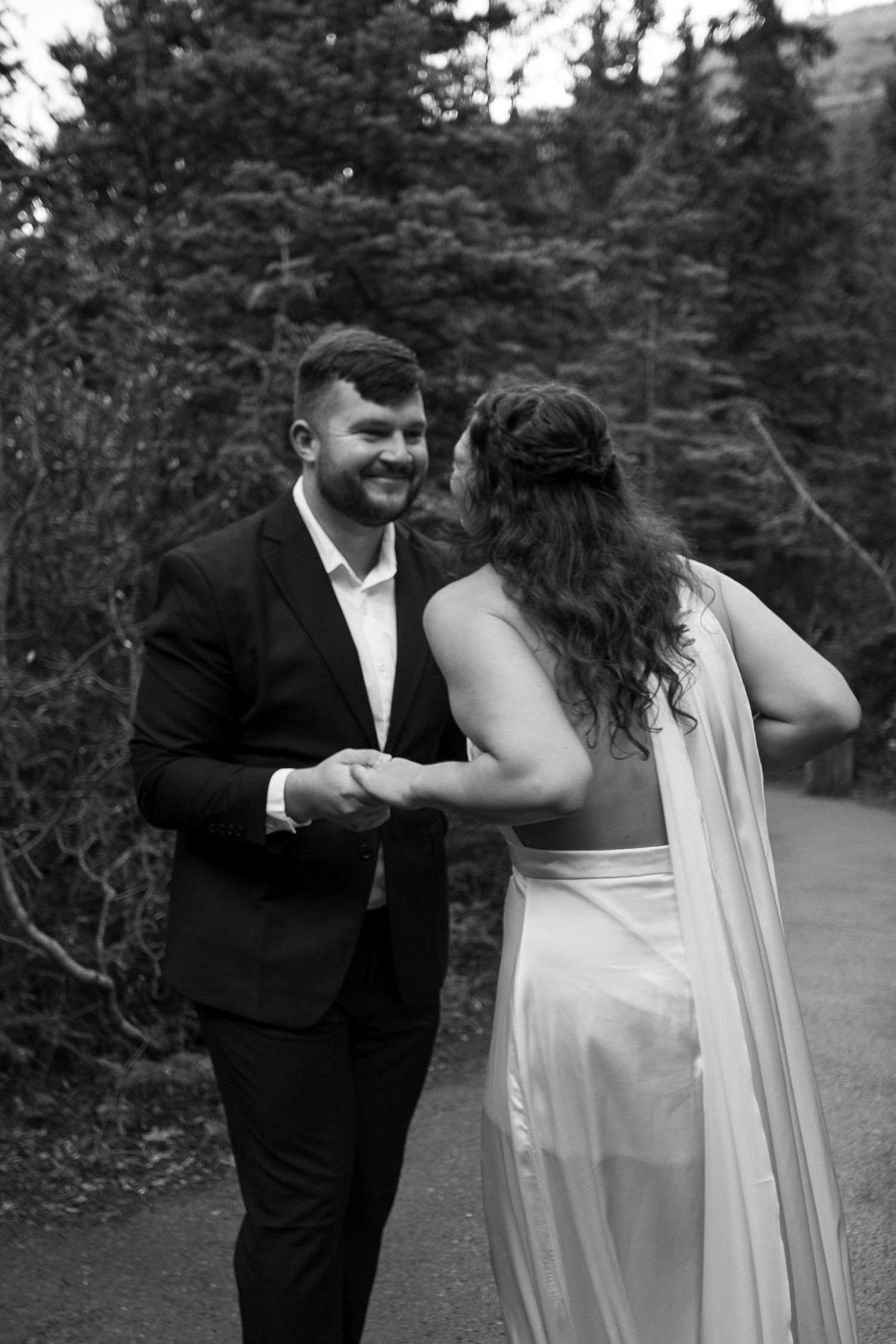Black and white photo of smiling groom in suit holding hands with bride in flowing gown on outdoor path.