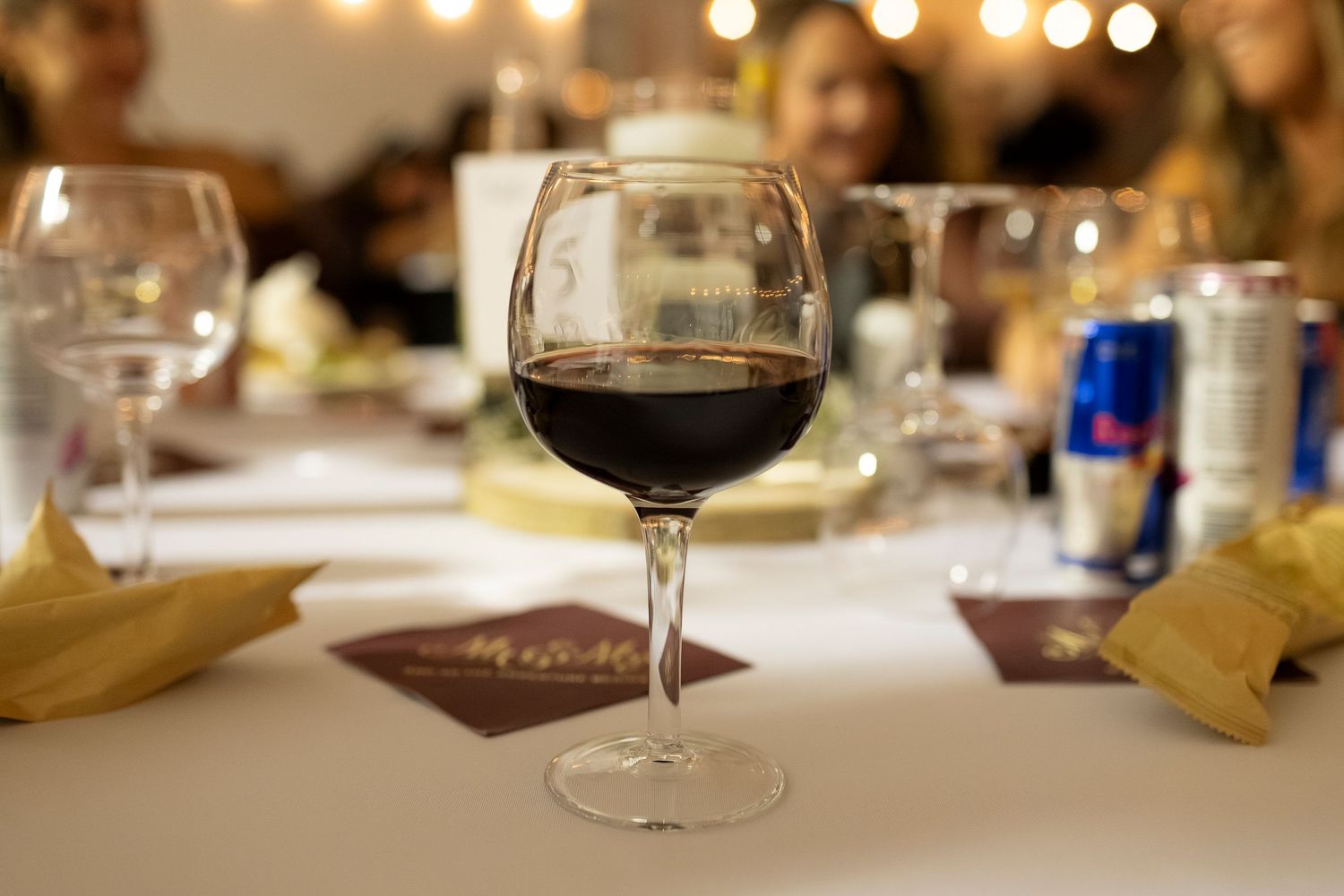 A glass of red wine on a white tablecloth at a dinner party with guests and a Red Bull can in the background.