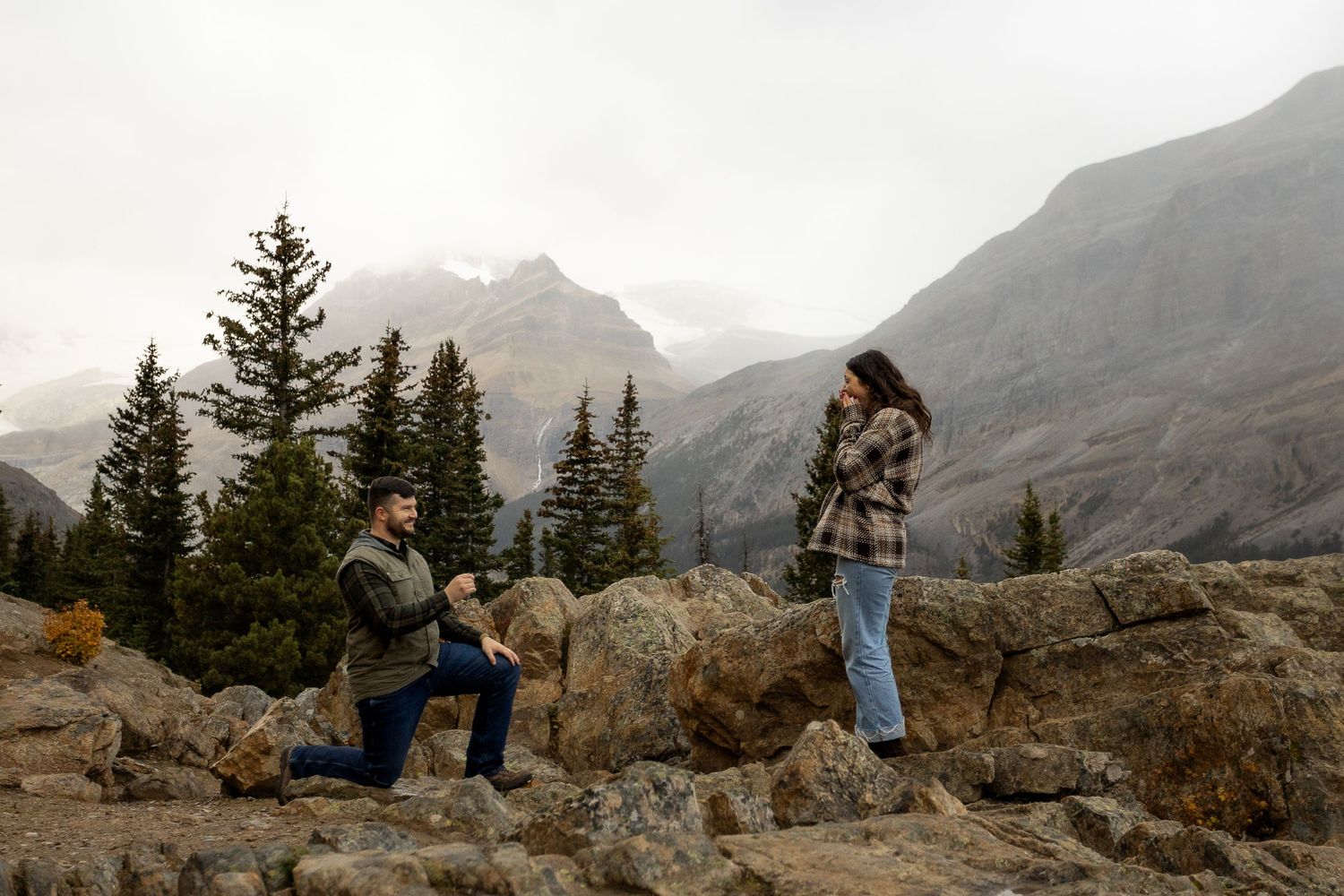 Man proposes on one knee to woman on rocky mountain terrain with evergreen trees and misty peaks in background.