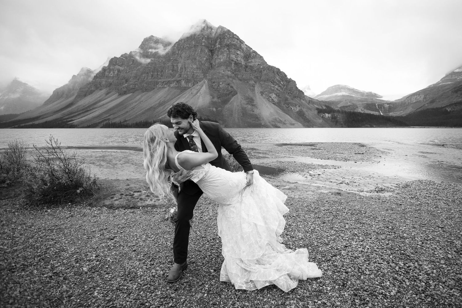 Black and white wedding photo of bride and groom sharing a romantic dip kiss by a mountain lake.
