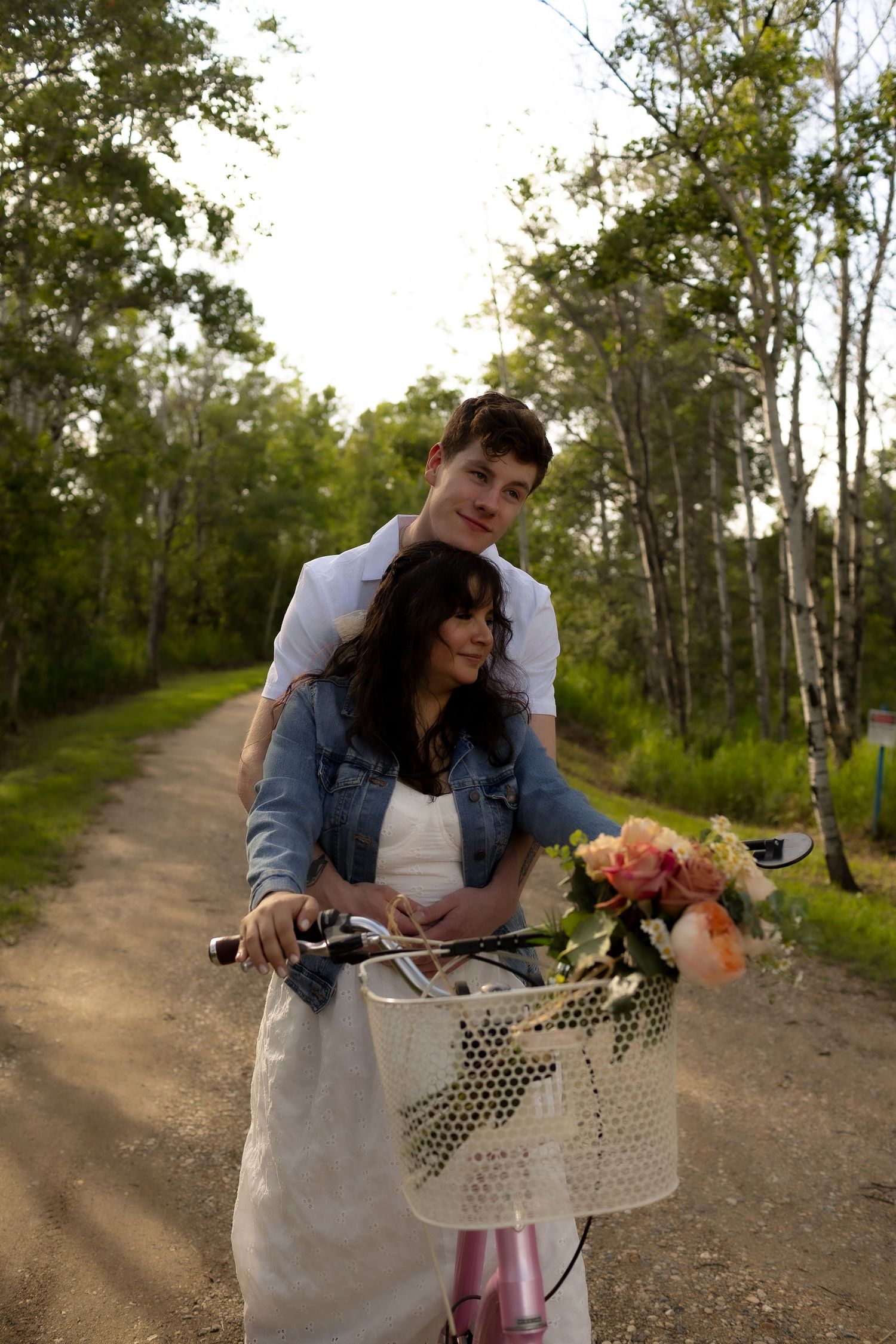 Couple shares a playful moment on a bicycle with flowers in the basket on a dirt path at the Lawrence House in Saskatoon, Saskatchewan.