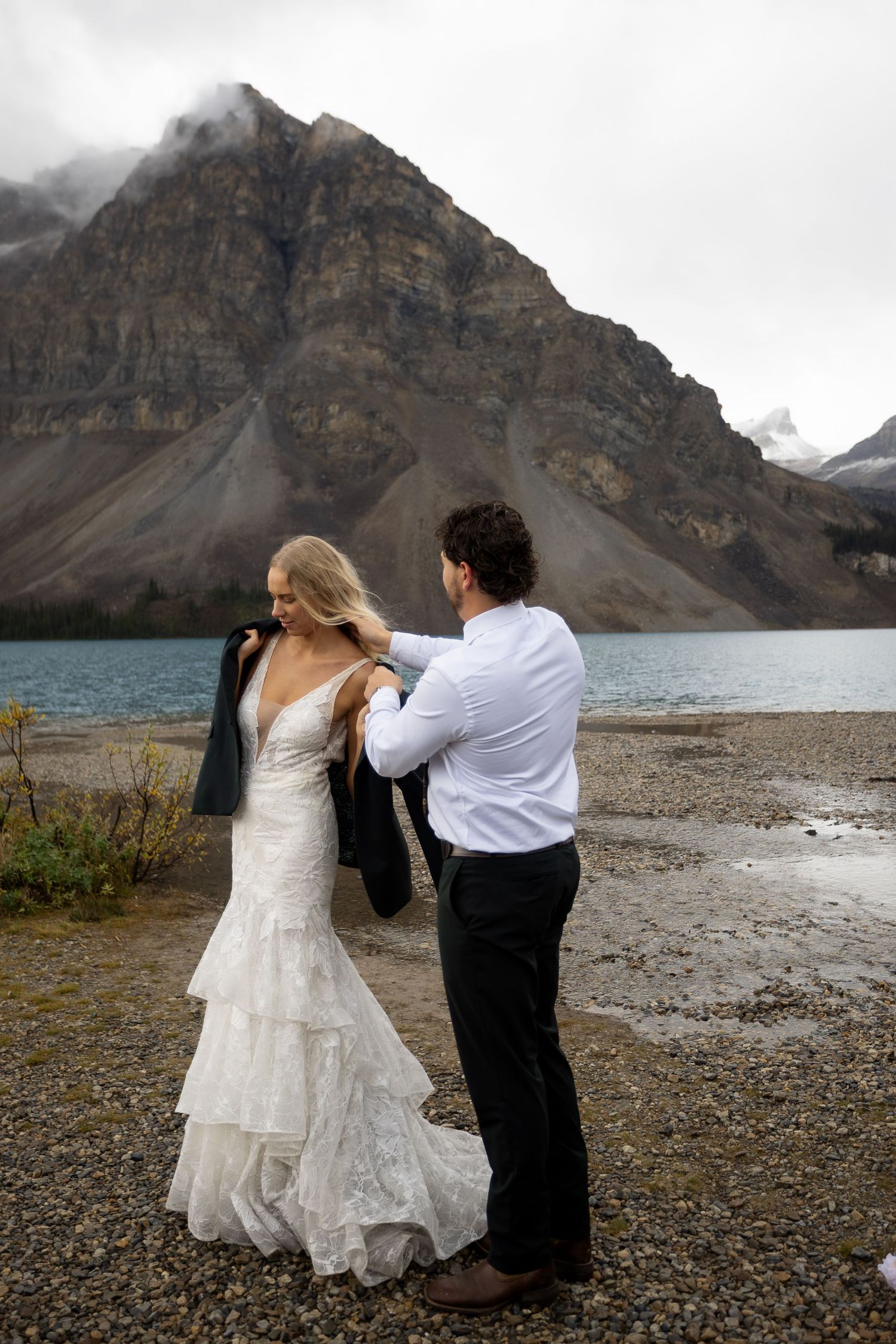 Groom placing jacket on bride in white lace wedding dress by a mountain lake with misty peaks in background.