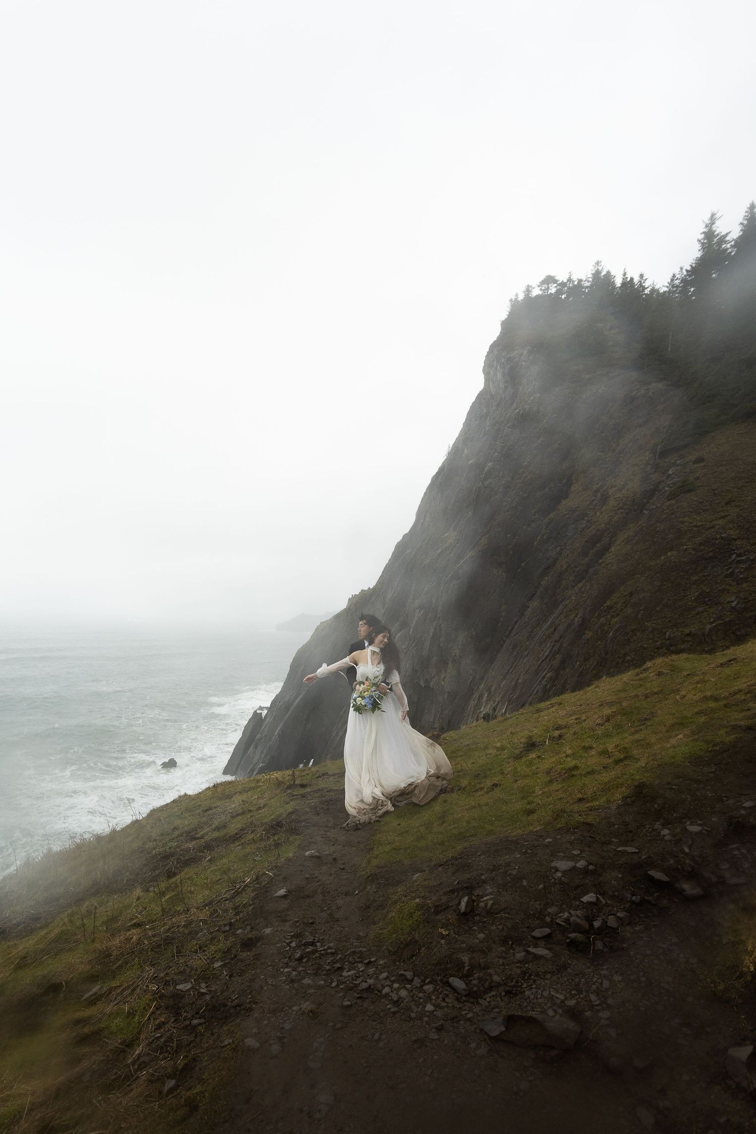 Bride in white gown holding bouquet stands on misty coastal cliff with ocean and rocky headland behind her.
