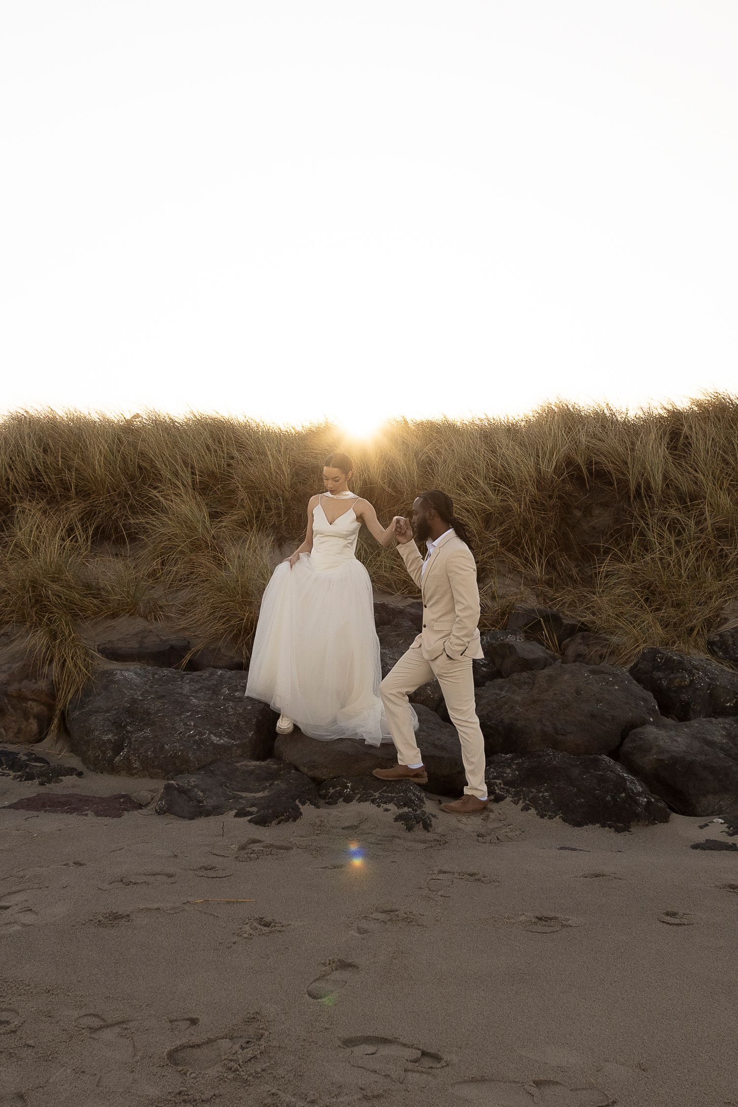 A couple walks along a sandy beach at golden hour with tall grass in the background.