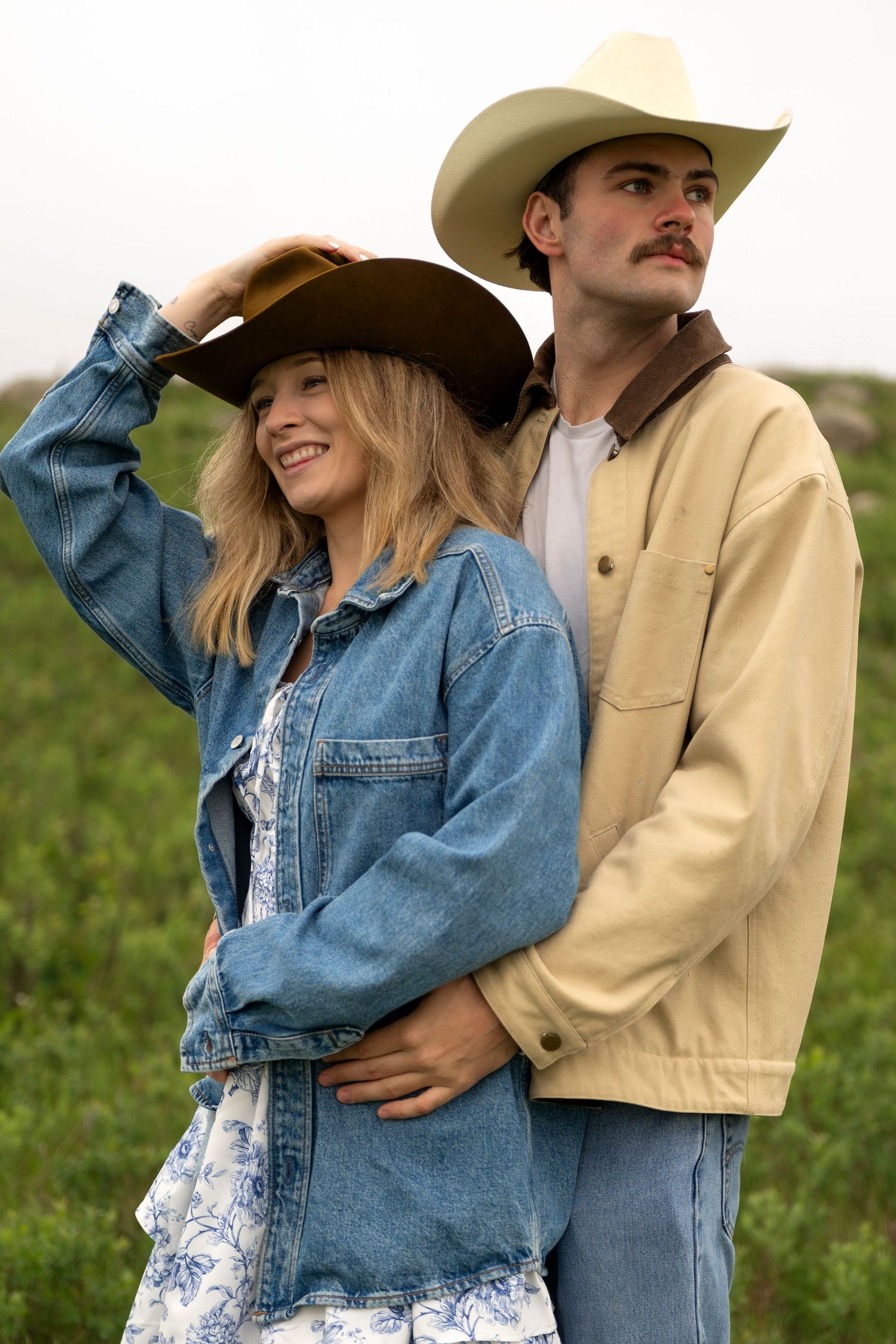 A smiling woman in a denim jacket and cowboy hat embraces a man in a tan jacket outdoors.