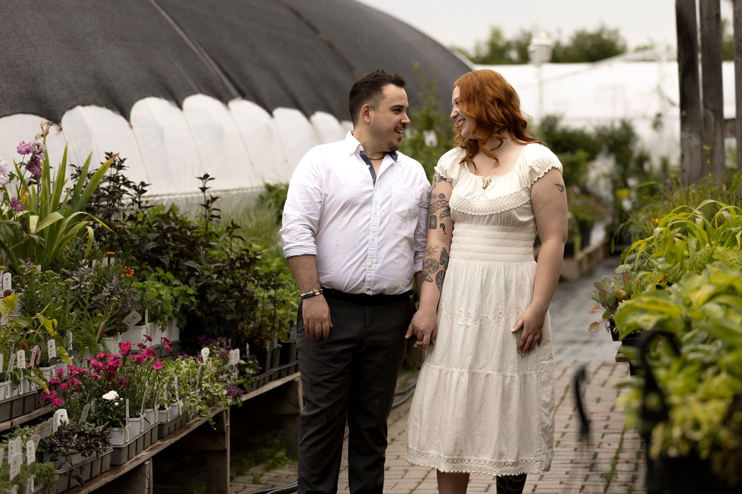 Couple in white attire holding hands among lush plants in a greenhouse garden setting.