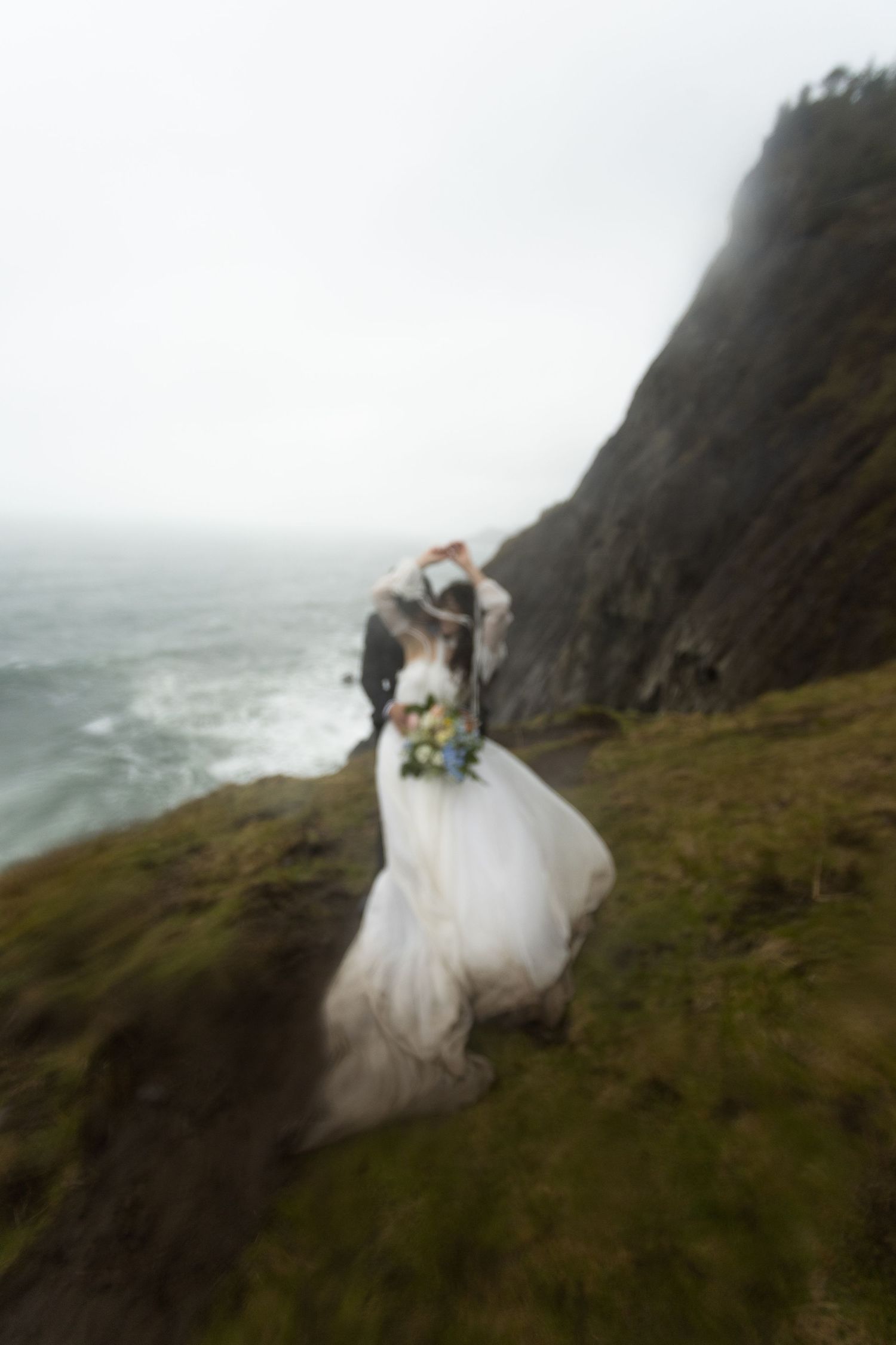 Couple in wedding attire embracing on a misty coastal cliff with ocean waves and dramatic rocky landscape.