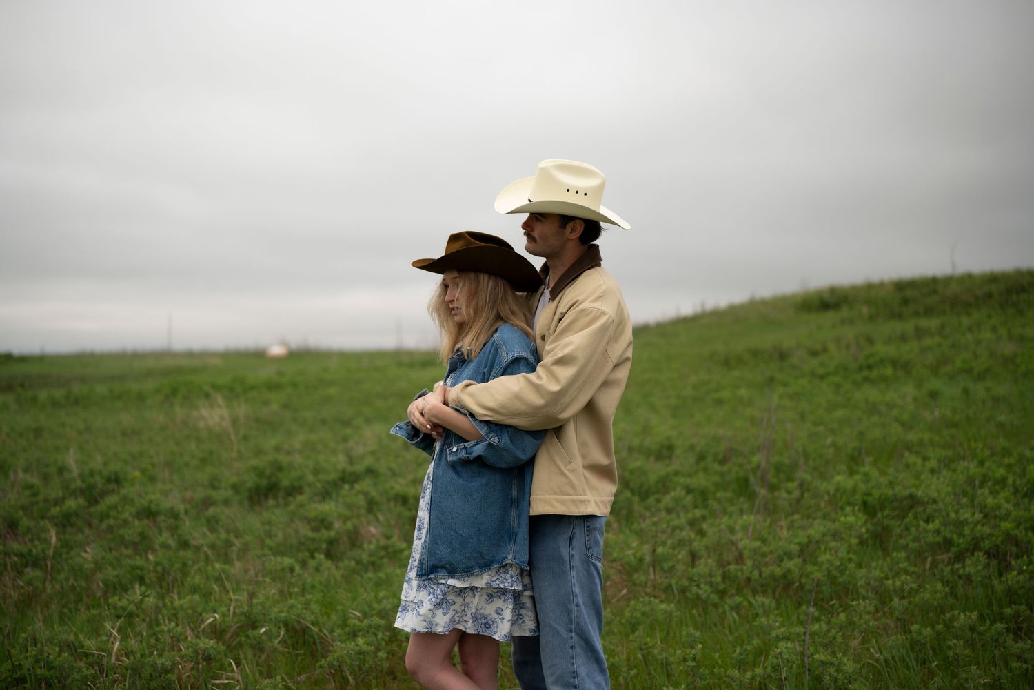 Couple in cowboy hats embracing in a green field under an overcast sky, wearing denim and beige outfits.