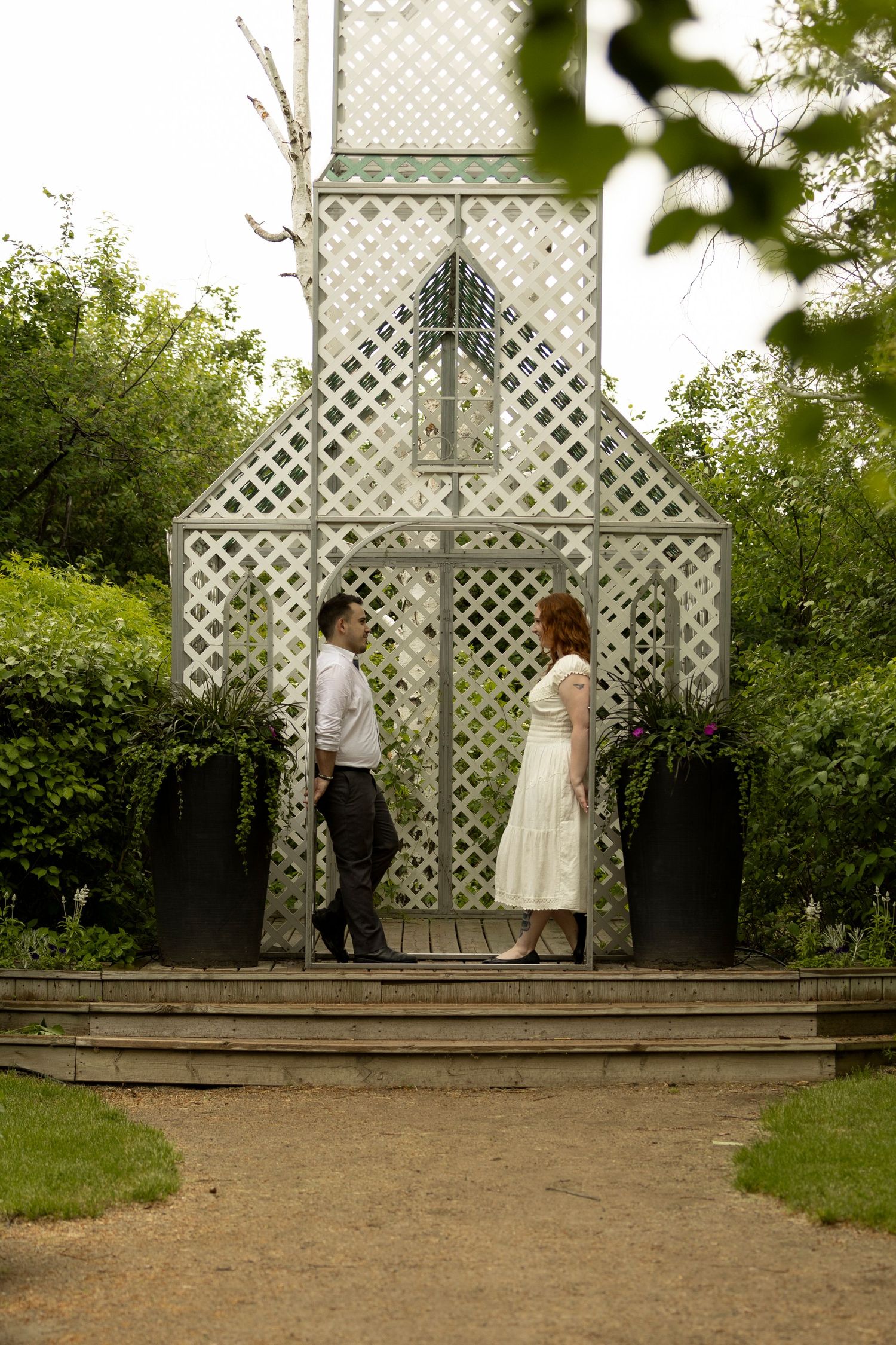 A couple stands facing each other at a white lattice garden chapel surrounded by lush greenery.