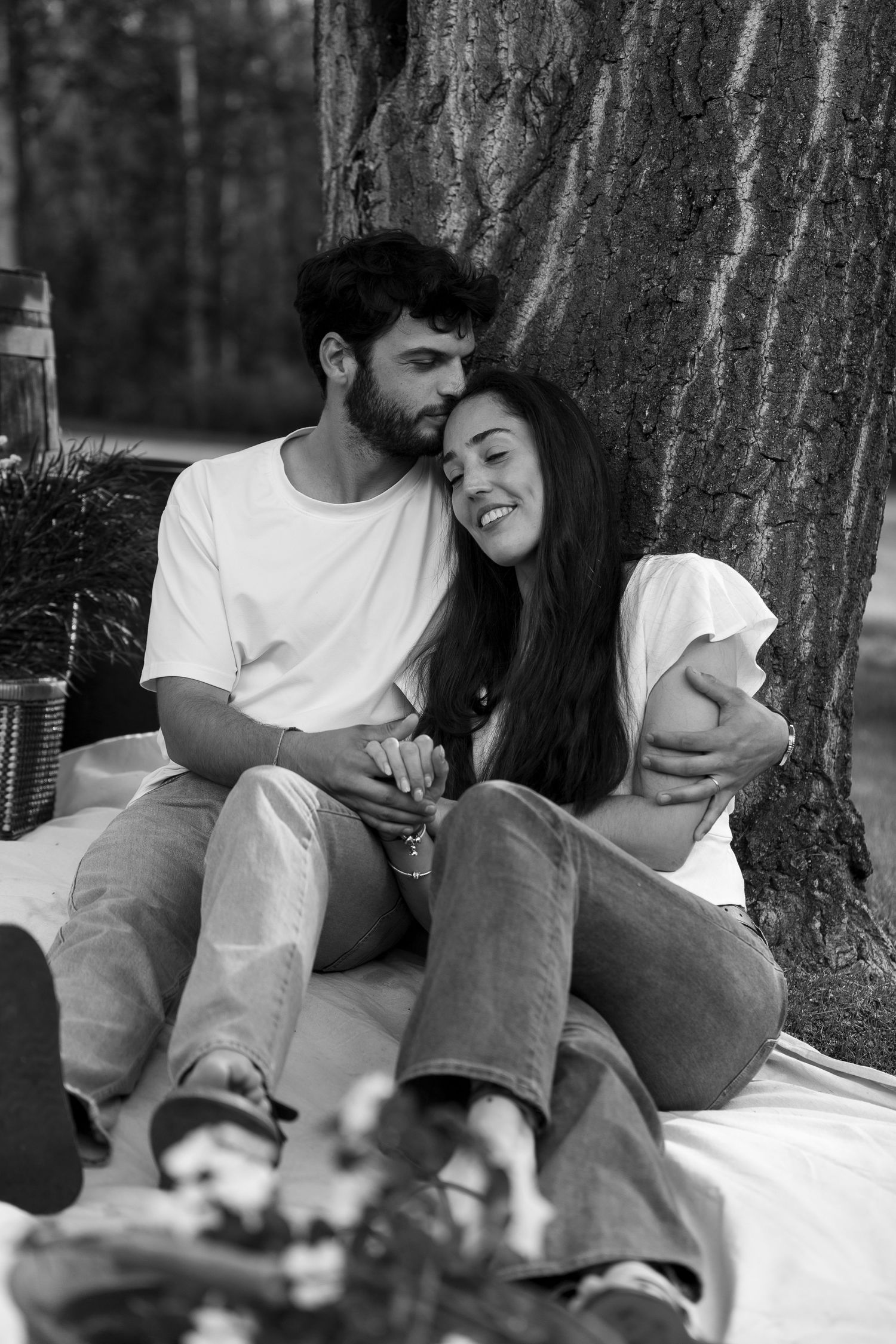 Black and white photo of a romantic couple sitting together under a tree, smiling and embracing during a picnic.